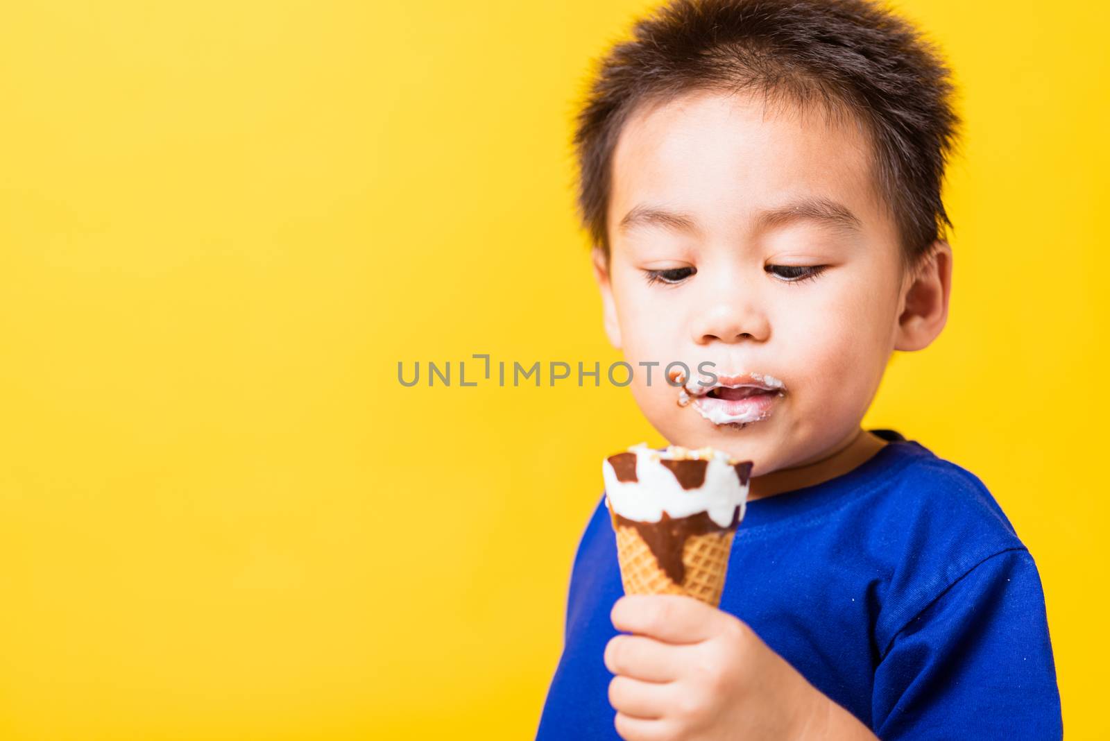 Happy portrait Asian child or kid cute little boy attractive laugh smile playing holds and eating sweet chocolate ice cream waffle cone, studio shot isolated on yellow background, summer concept