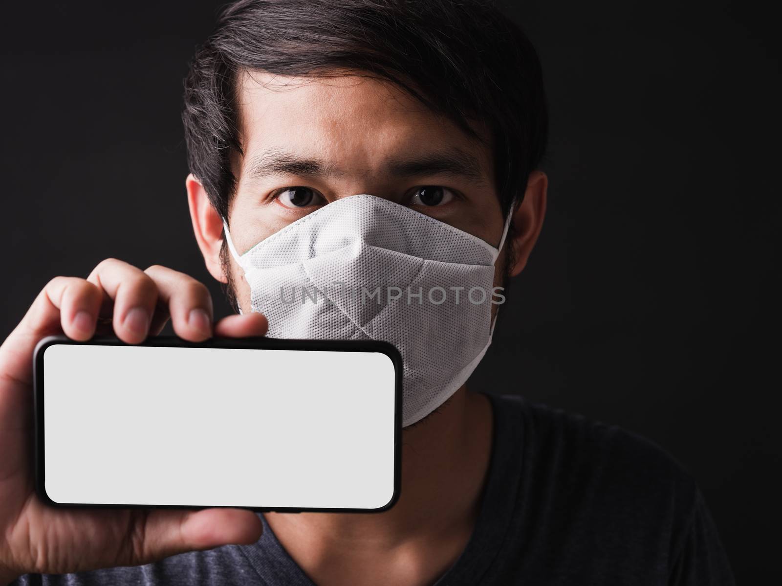 Closeup Asian handsome man wearing protective face mask fear in eye holding show mobile phone blank screen near face, hygiene prevention COVID-19 or coronavirus protection concept, black background