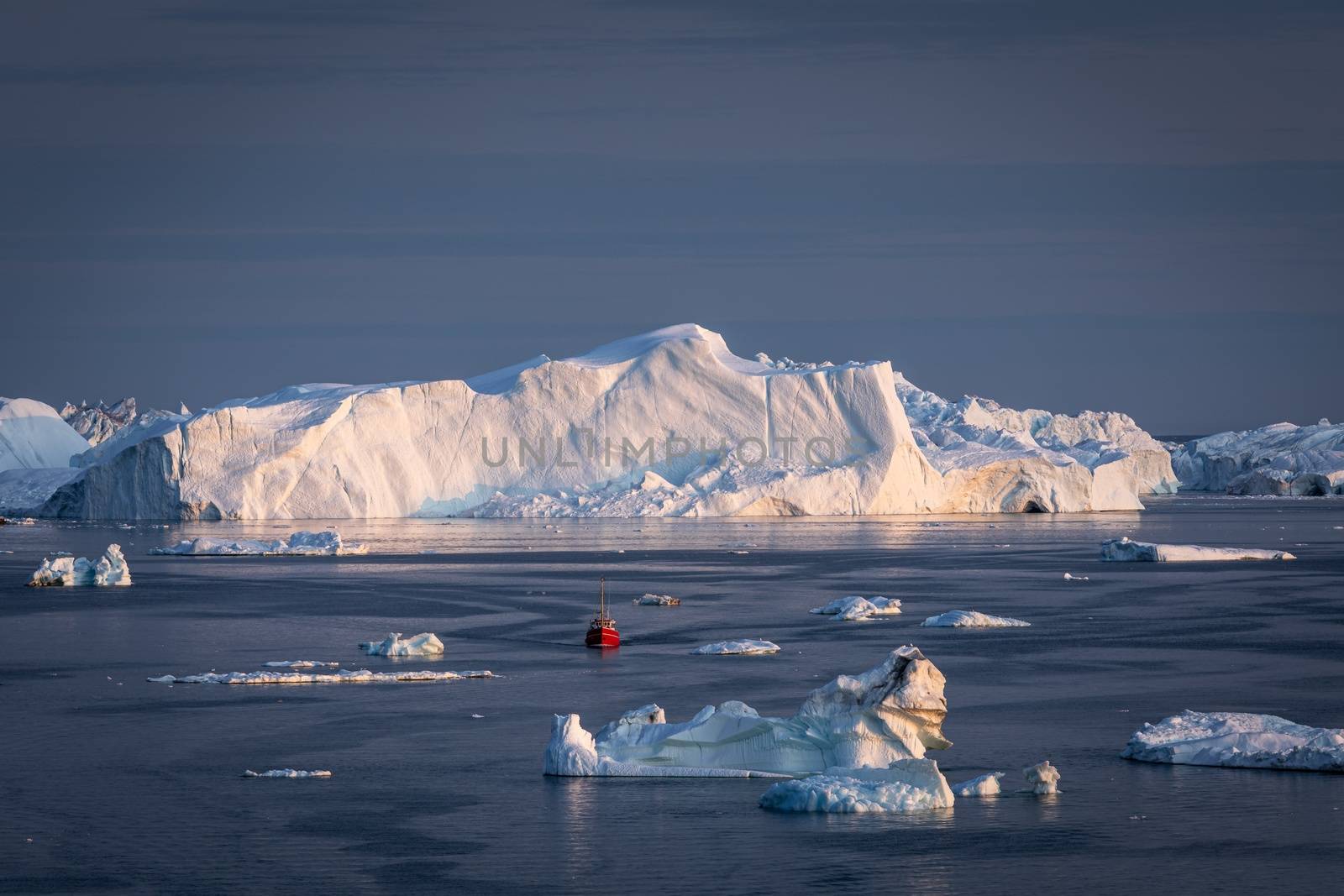 Little red sailboat cruising among floating icebergs in Disko Bay glacier during midnight sun season of polar summer. Ilulissat, Greenland. by bhavik_jagani