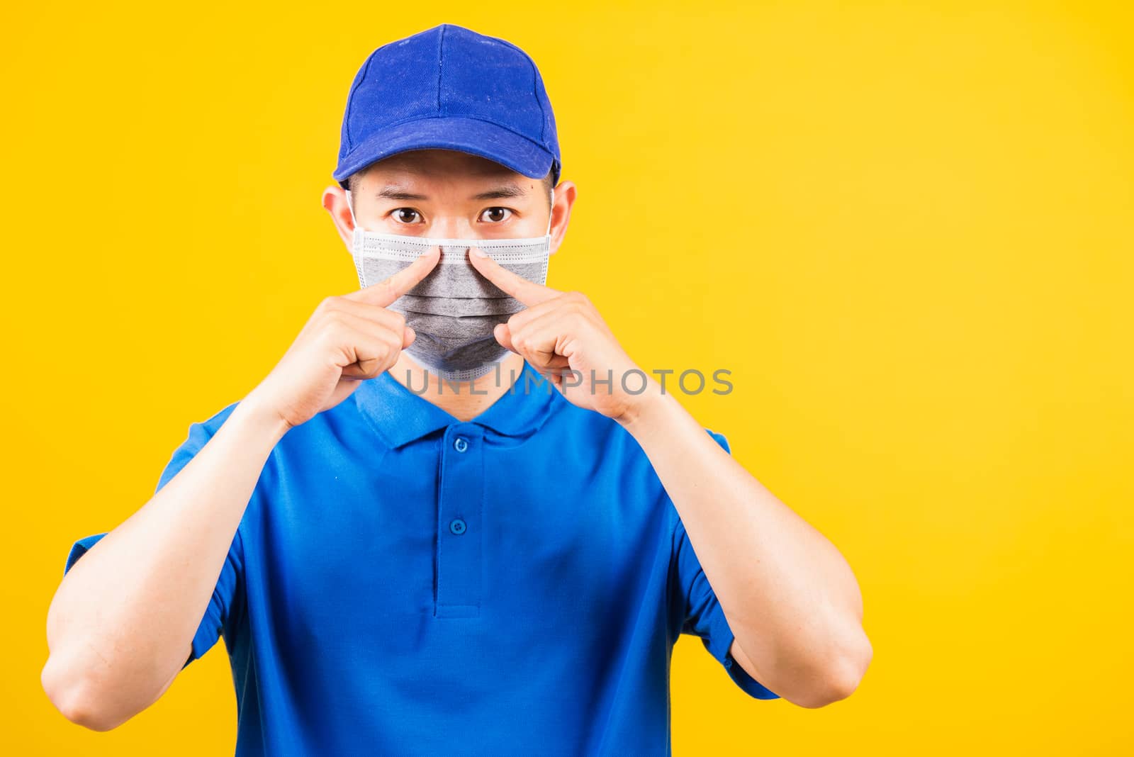 Asian young delivery worker man in blue t-shirt and cap uniform wearing face mask protective for service customer under quarantine pandemic coronavirus COVID-19, studio shot isolated yellow background