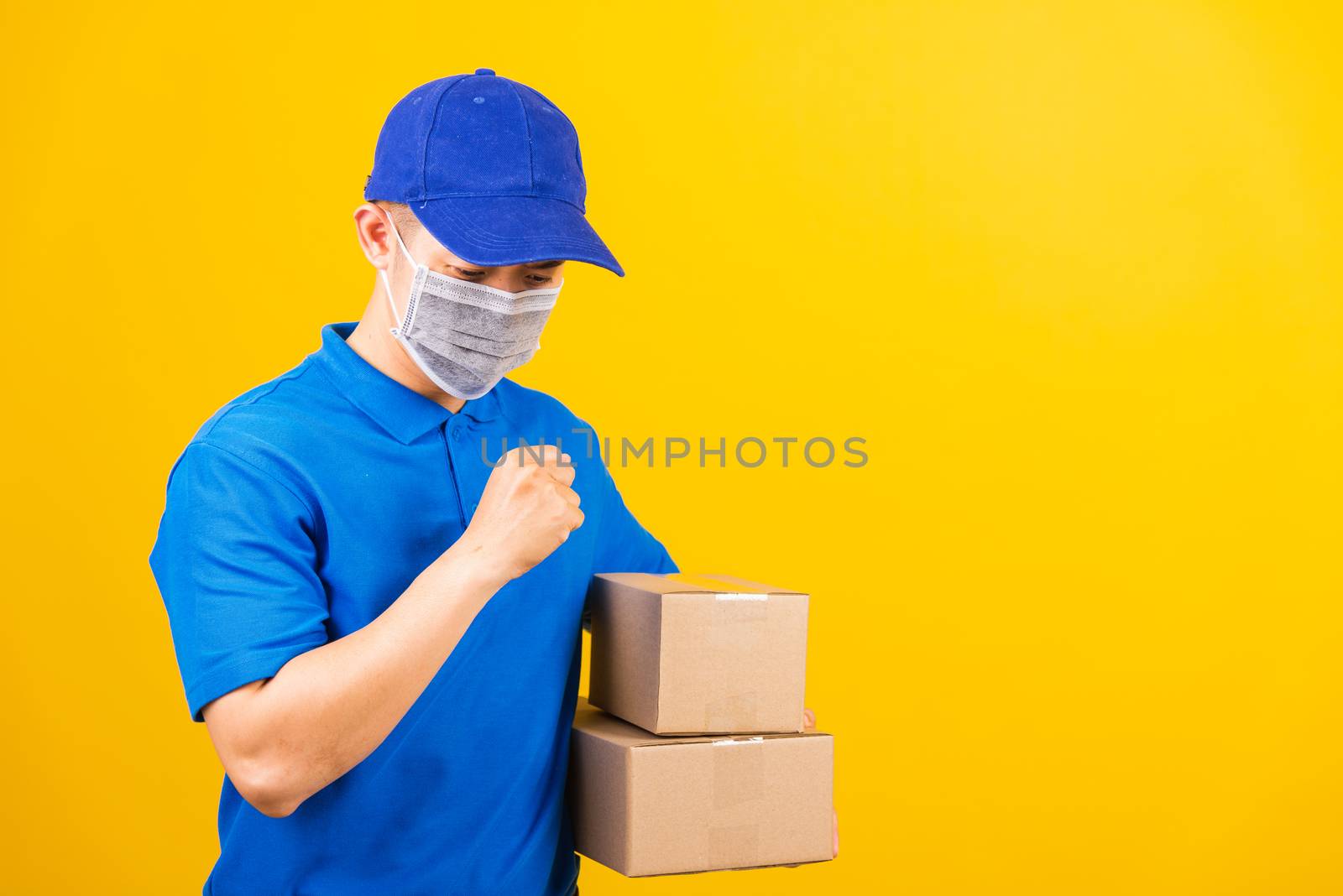Asian young delivery worker man in blue t-shirt and cap uniform wearing face mask protective he sneezing during service customer under coronavirus or COVID-19, studio shot isolated yellow background