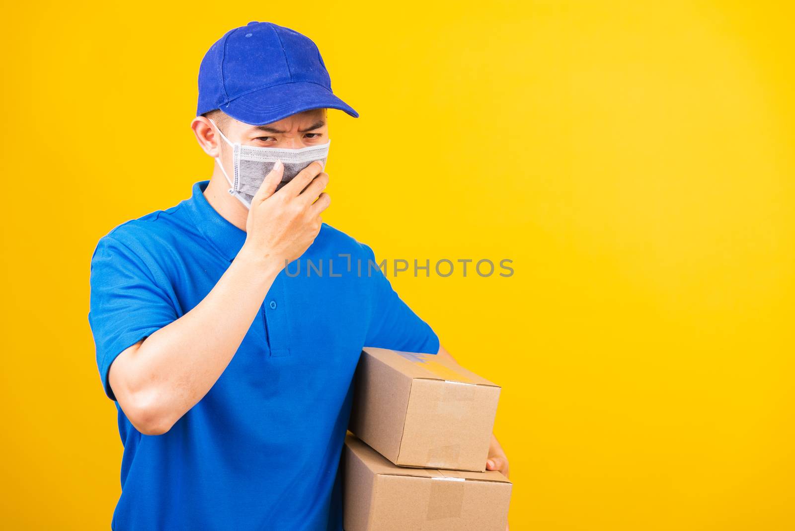 Asian young delivery worker man in blue t-shirt and cap uniform wearing face mask protective he sneezing during service customer under coronavirus or COVID-19, studio shot isolated yellow background