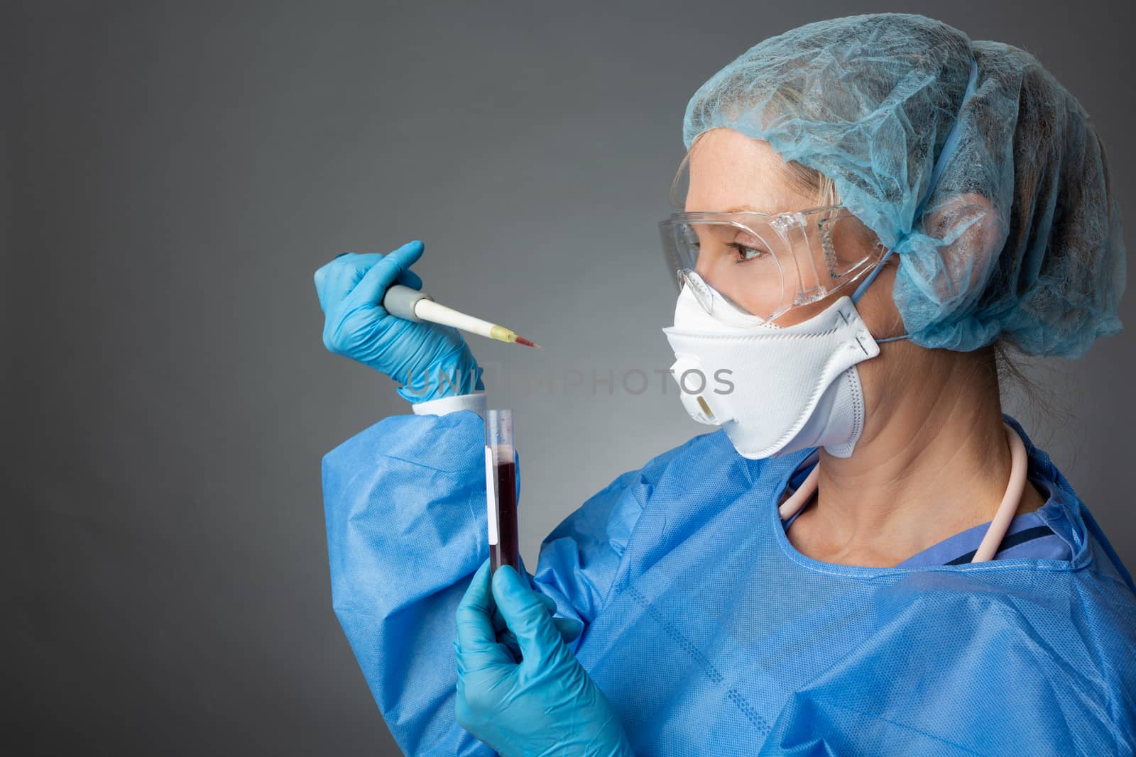 Laboratory pathologist healthcare worker holding a pipette blood sample for analysis.  She is wearing full protective PPE including gloves, apron, goggles, respirator mask