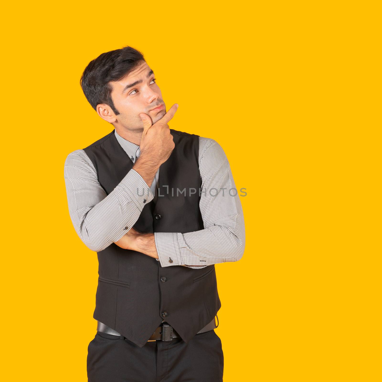 A young businessman in a gray shirt and a black vest with his hands held his chin, contemplating the project being started. Concept of investment in business. Portrait on yellow background in studio.