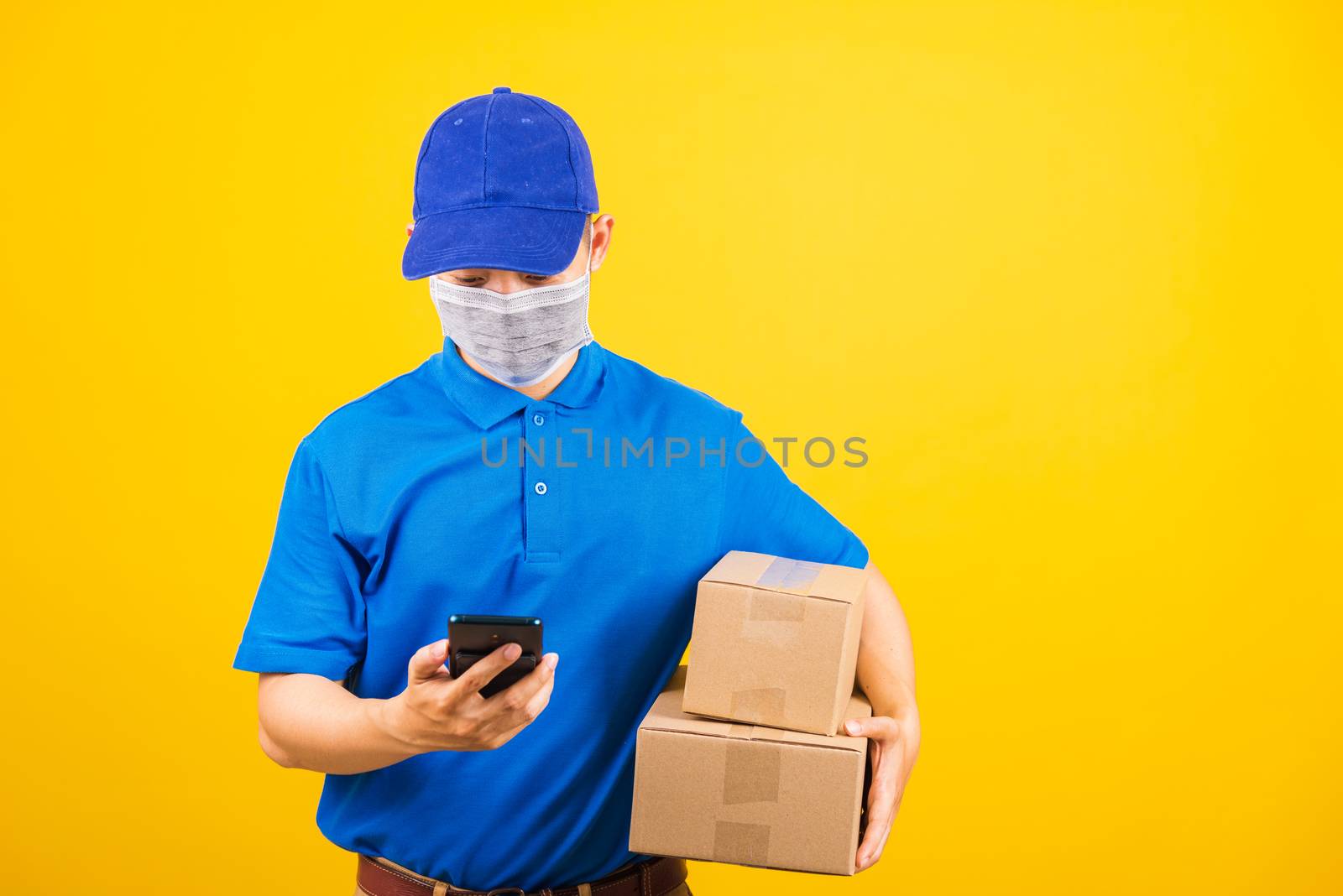 Asian young delivery worker man t-shirt and cap uniform wearing face mask protective check location of smart mobile phone under coronavirus or COVID-19, studio shot isolated yellow background