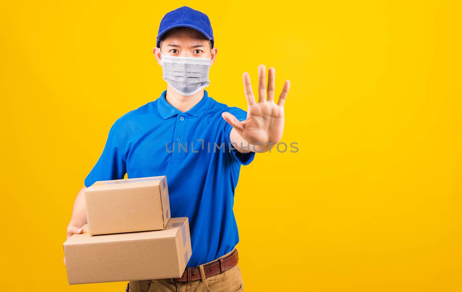 Asian young delivery worker man in blue t-shirt and cap uniform wearing face mask protective hold boxes he raise hand to stop sign under coronavirus or COVID-19, studio shot isolated yellow background