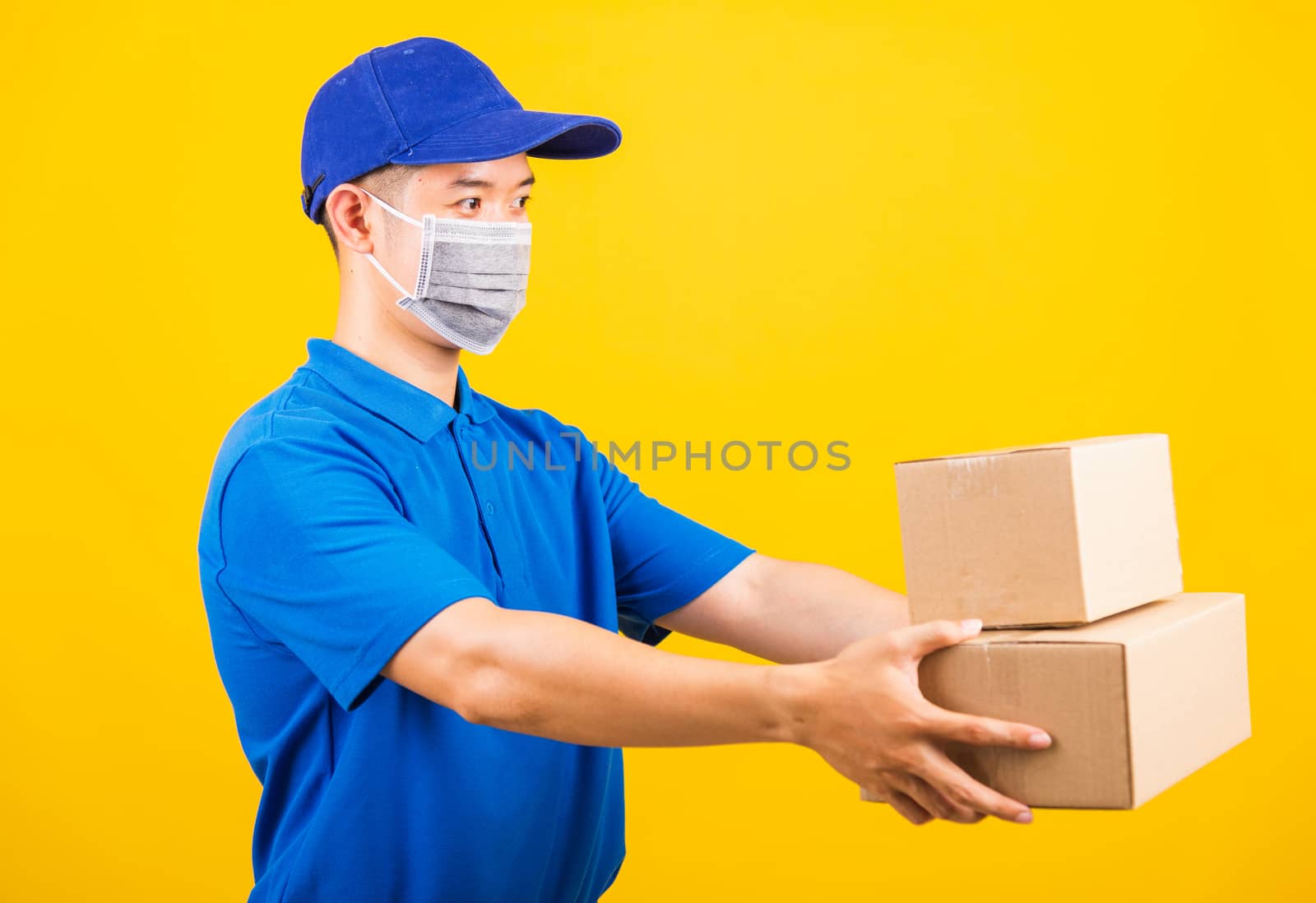 Side of Asian young delivery worker man in blue t-shirt and cap uniform wearing face mask protective giving cardboard boxes under coronavirus or COVID-19, studio shot isolated yellow background