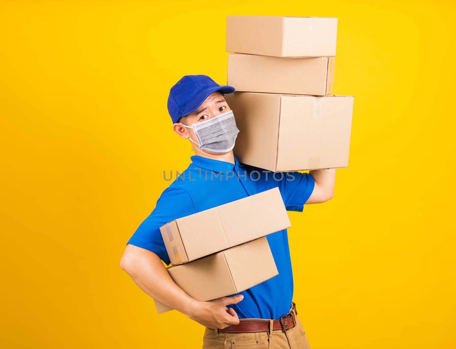 Asian young delivery worker man in blue t-shirt and cap uniform wearing face mask protective lifting stack a lot of boxes, under coronavirus or COVID-19, studio shot isolated yellow background