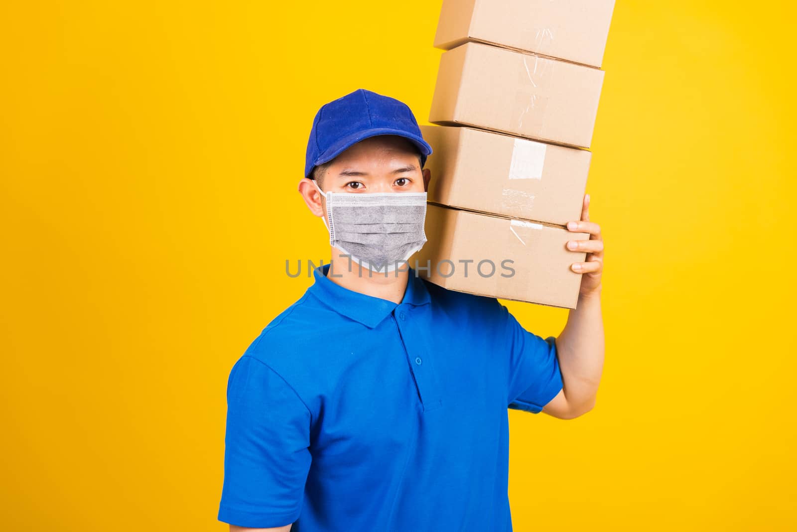 Asian young delivery worker man in blue t-shirt and cap uniform wearing face mask protective lifting stack a lot of boxes, under coronavirus or COVID-19, studio shot isolated yellow background