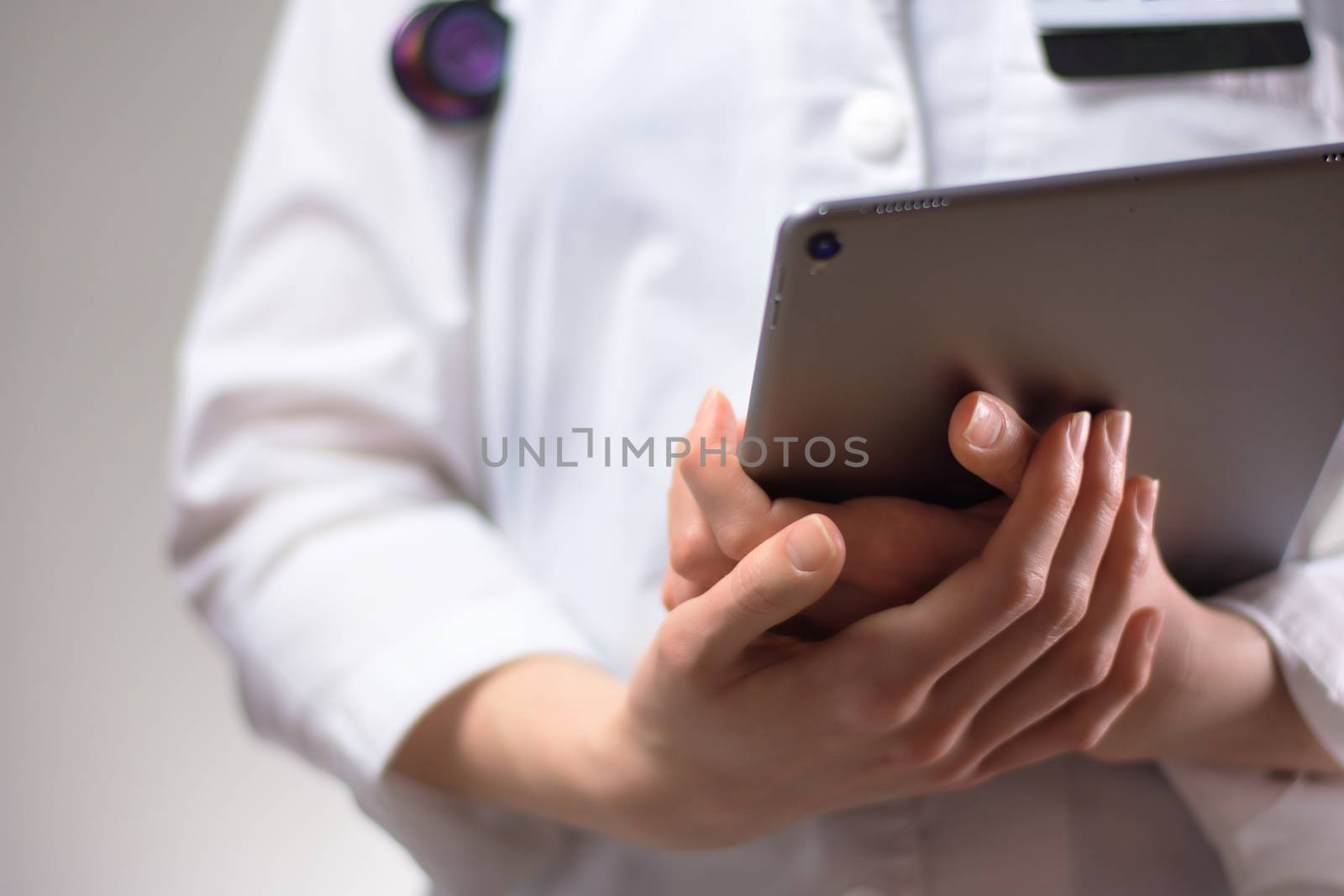 Tablet in the hands of healthcare professional up close. White coat, stethoscope, and badge visible in background. Hands of nurse practitioner or PA using technology in medicine for patients by jmac23