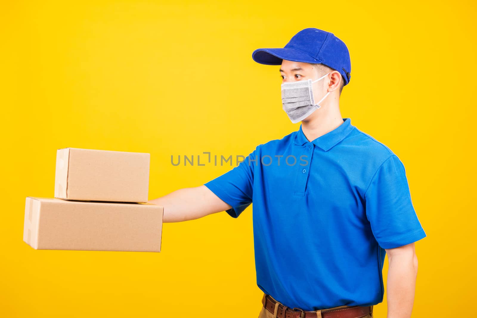 Side of Asian young delivery worker man in blue t-shirt and cap uniform wearing face mask protective giving cardboard boxes under coronavirus or COVID-19, studio shot isolated yellow background