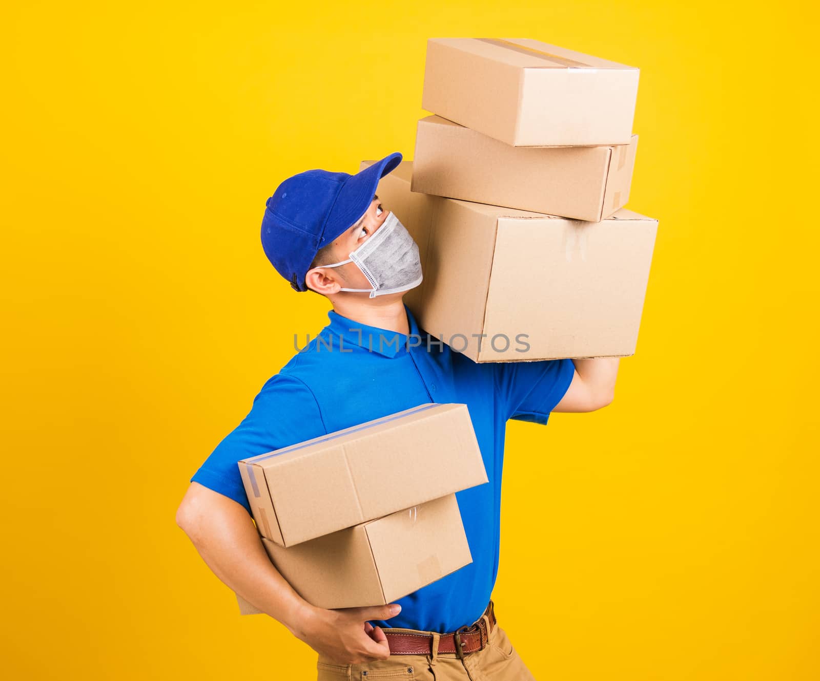 Asian young delivery worker man in blue t-shirt and cap uniform wearing face mask protective lifting stack a lot of boxes, under coronavirus or COVID-19, studio shot isolated yellow background