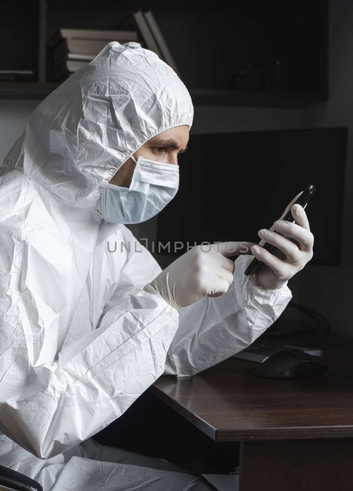 Man in protective suit, medical mask and rubber gloves sits at home and using a phone for chatting with a friends or business partners during quarantine. Man at remote work in a pandemic covid. Coronavirus