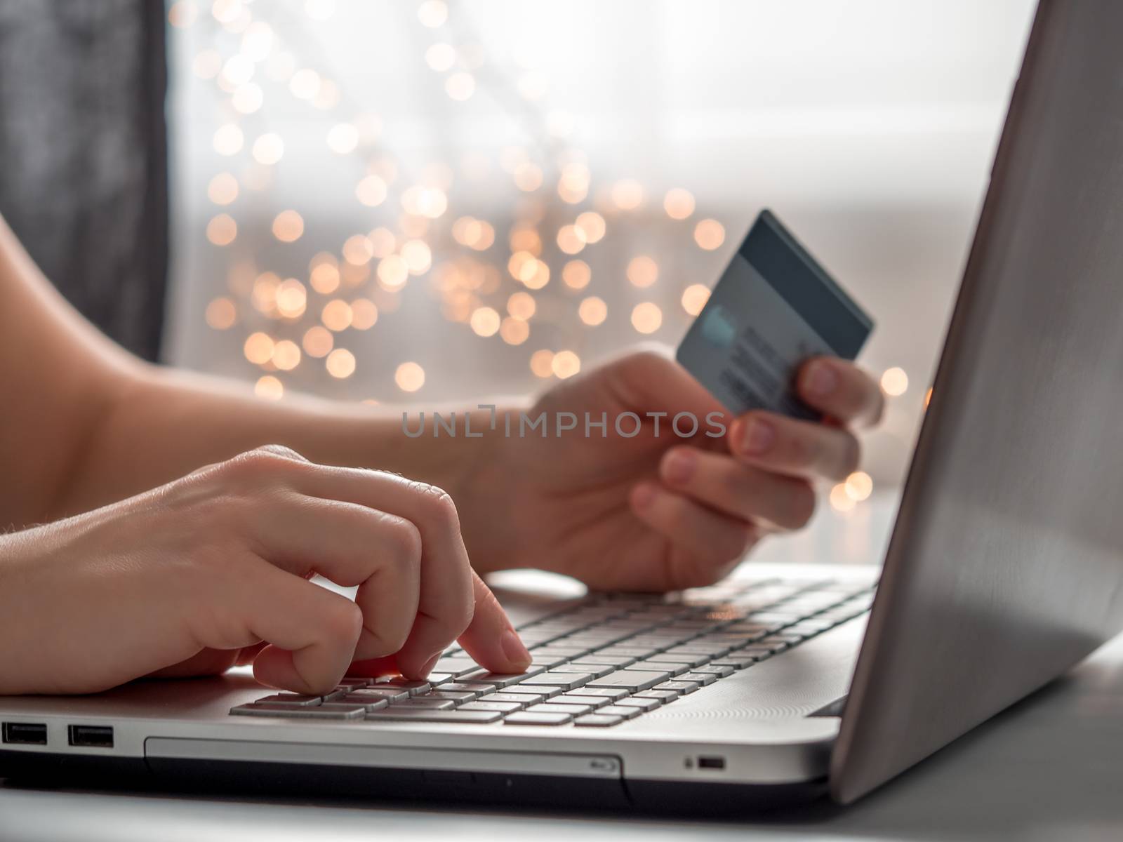 Cristmas online shopping soncept. Close-up woman's hands holding credit card and inputting card information using laptop keyboard for online shopping with festive lighting chain bokeh.