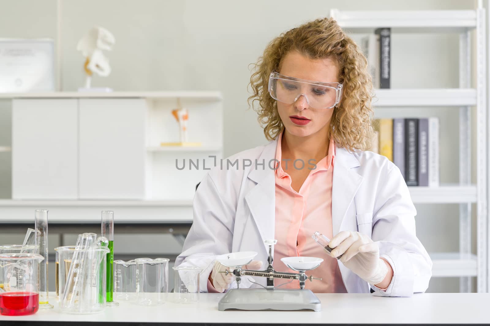 A young blonde scientist is measuring the chemicals for an experiment. Working atmosphere in chemical laboratory. Test tubes and beakers filled with chemicals on the table.