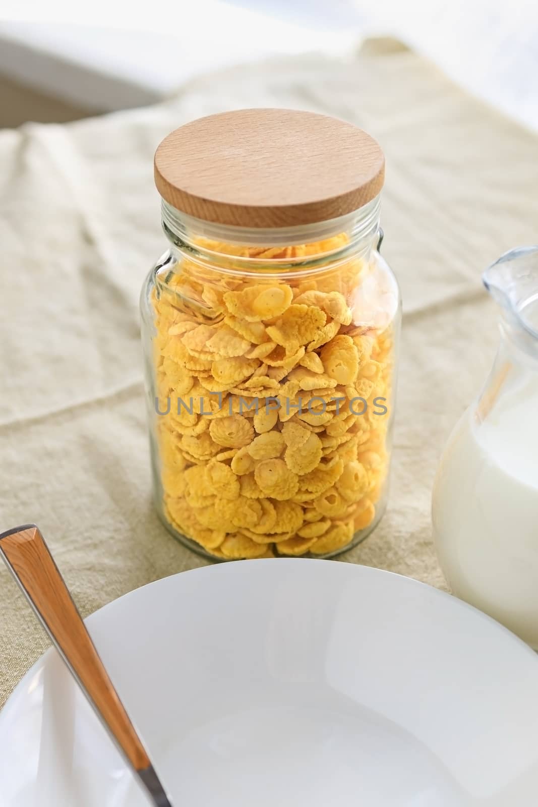 Cornflakes in a jar, next to a transparent jug of milk, an empty plate with a spoon on a plain rough tablecloth. View from above. by Tanacha