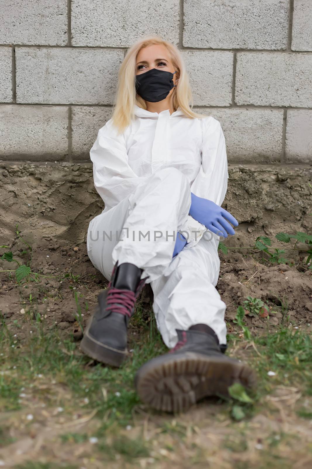 Woman in black mask, white protective suit and gloves sitting against wall