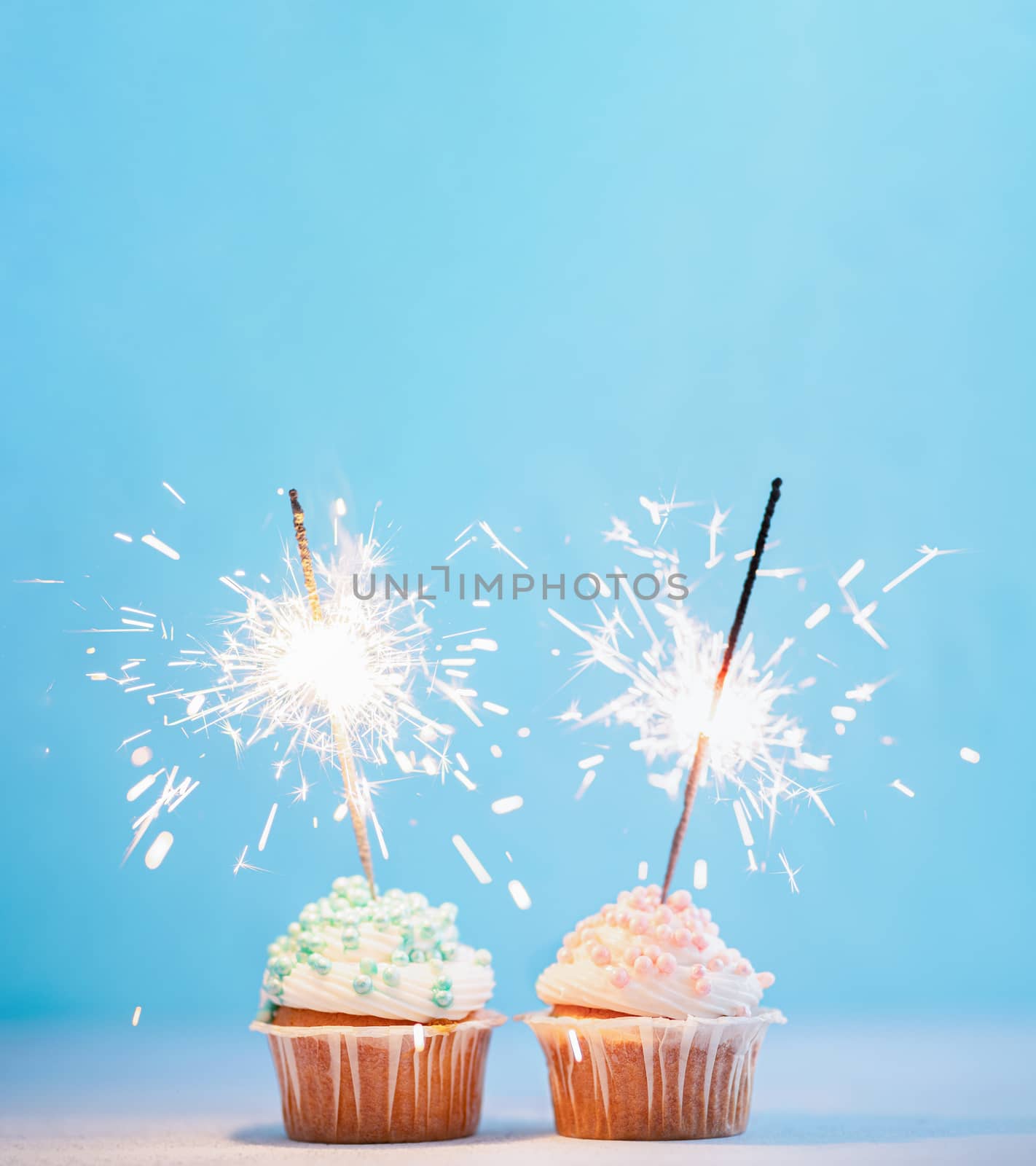 Two cupcakes decorated with colorful sprinkles and sparklers. Festive cupcakes with blue and pink sprinkles on blue background with copy space top for text or design