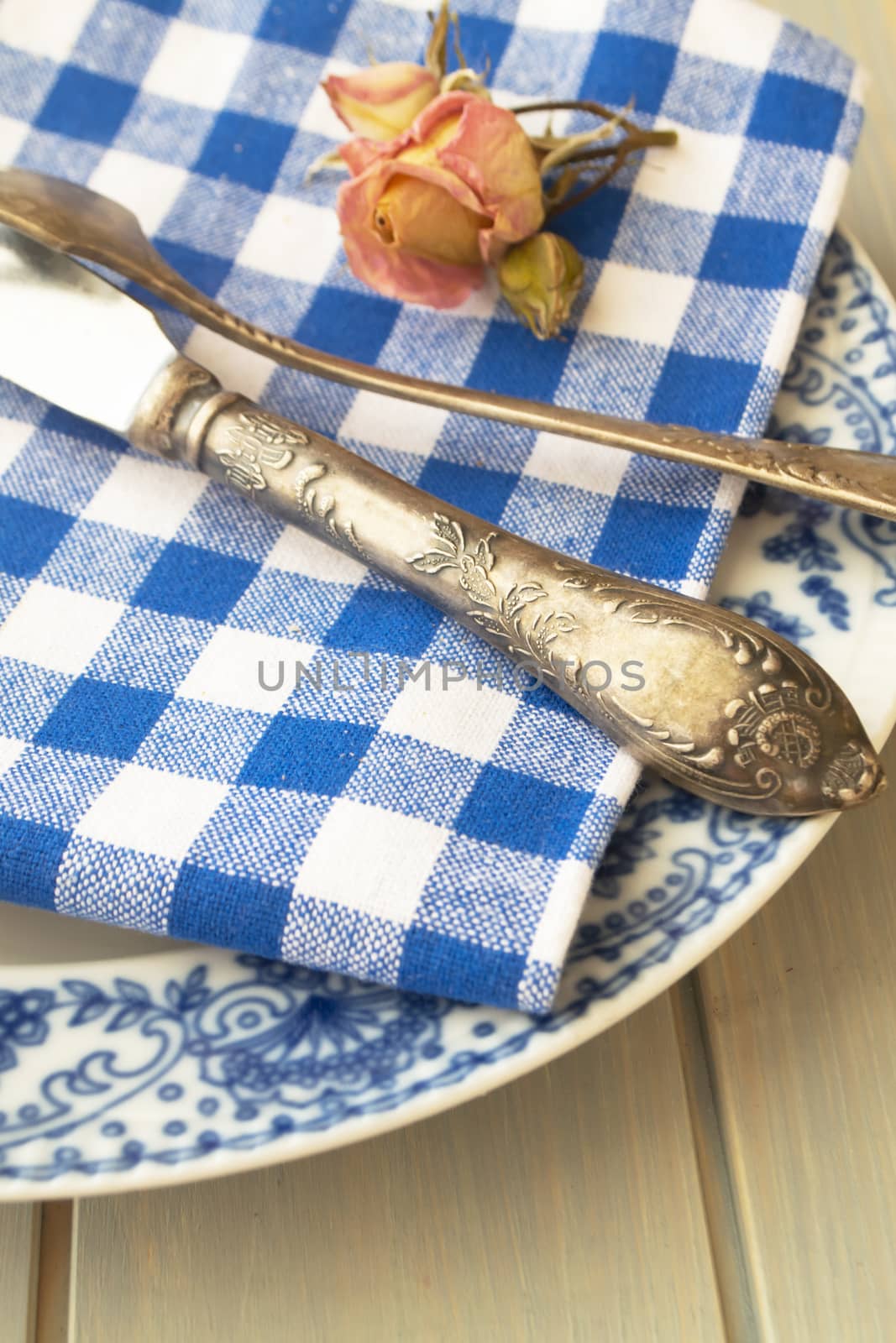 Silver fork and knife with an empty vintage plate on a blue napkin, with dry pink rose