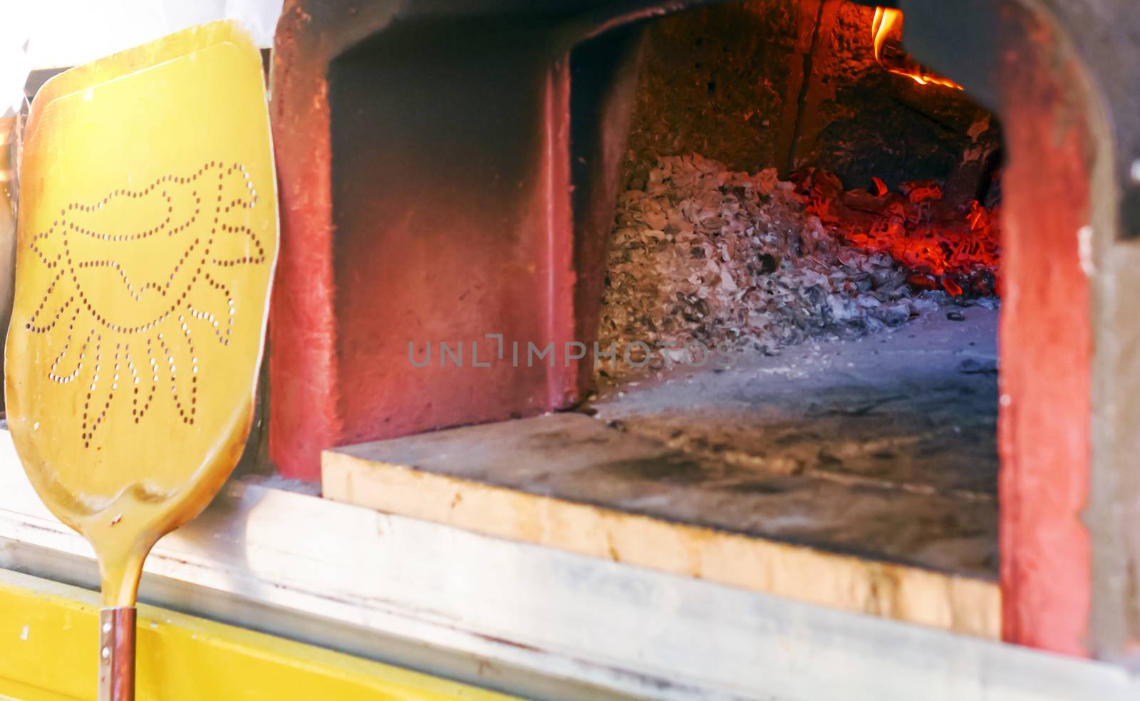 wood oven for baking pizza with ashes, burning embers and fire. Leaning against the oven there is a yellow shovel to take the pizza from the oven once it is ready