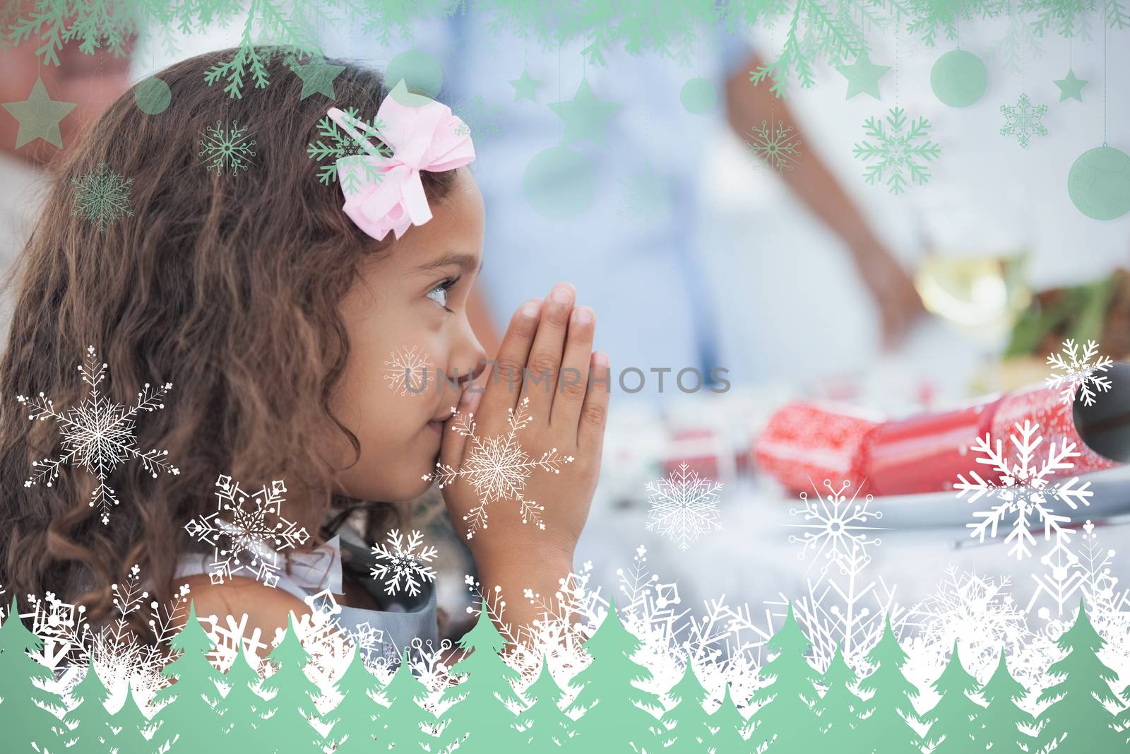 Little girl sitting praying at table against snowflakes and fir trees in green