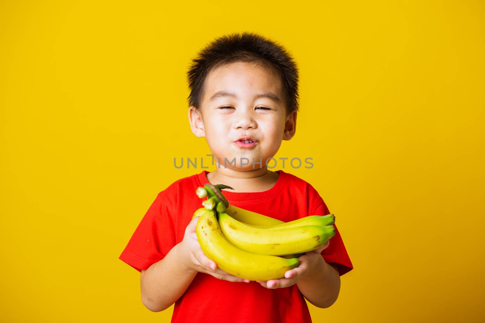 Happy portrait Asian child or kid cute little boy attractive smile wearing red t-shirt playing holds bananas comb fruit, studio shot isolated on yellow background