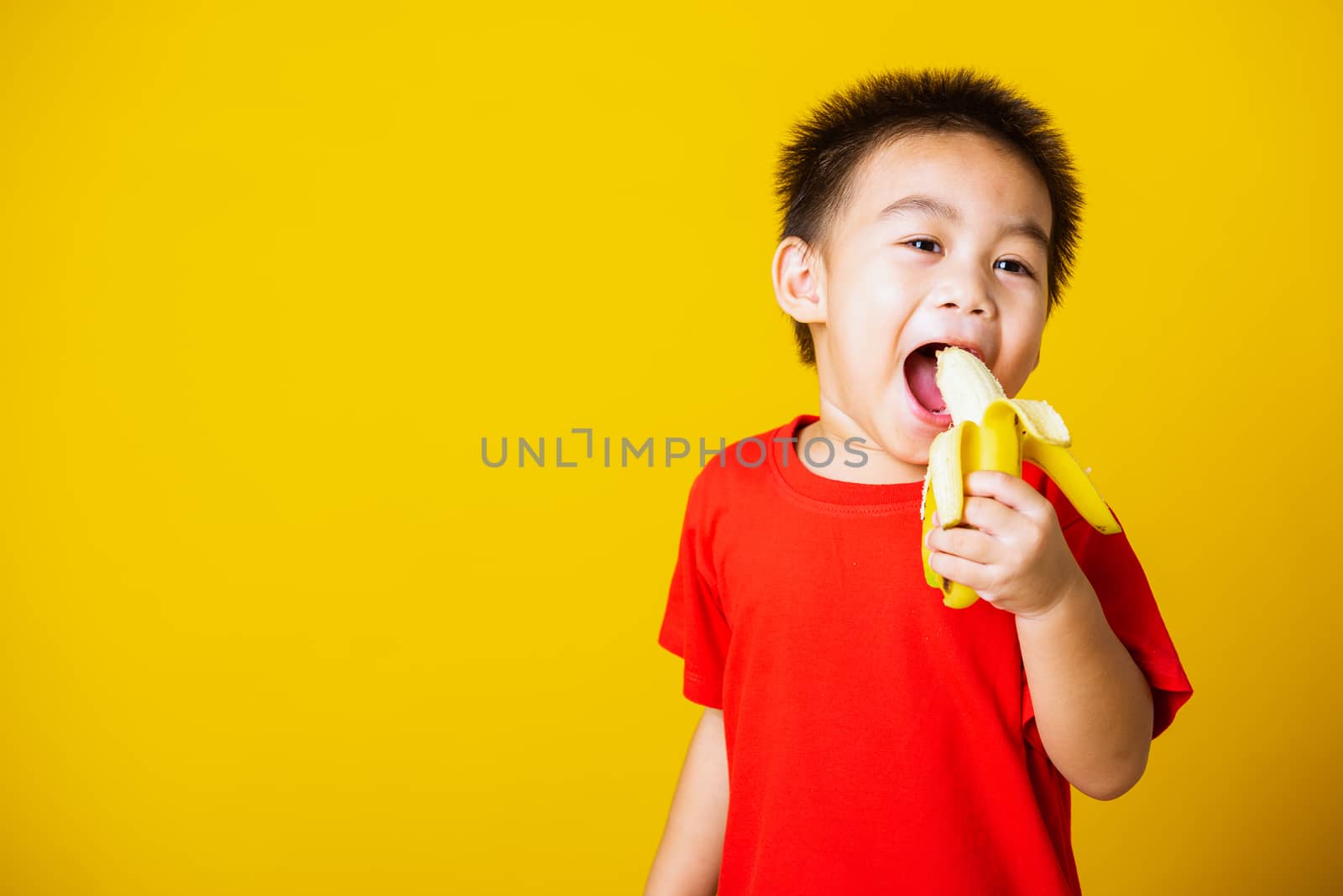 Happy portrait Asian child or kid cute little boy attractive smile wearing red t-shirt playing holds peeled banana for eating, studio shot isolated on yellow background