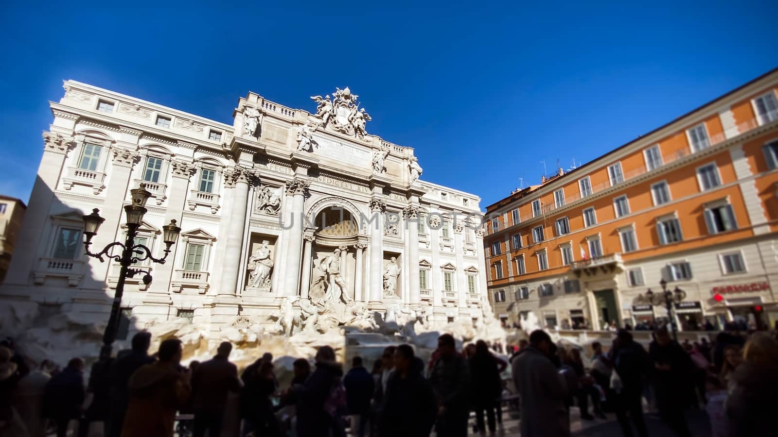 The marble facade of the Trevi Fountain illuminated by the sun with tourists walking on a sunny winter day in Rome.