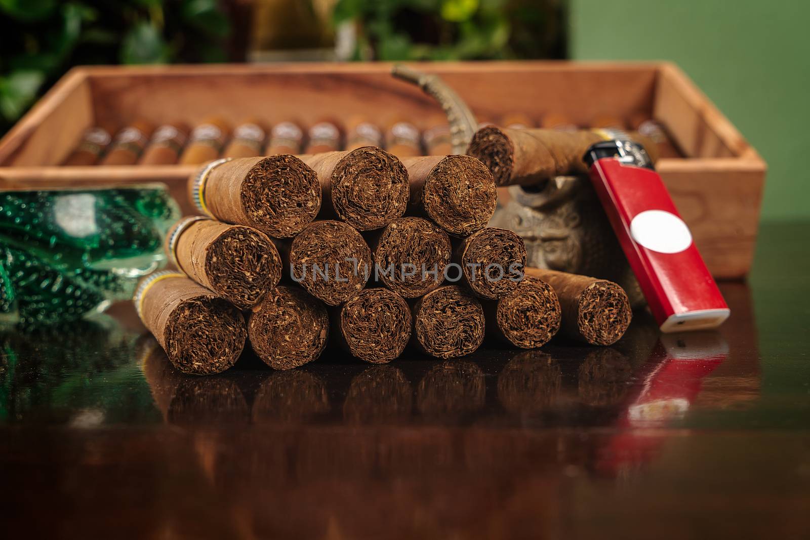 A group of cigars, a lighter, two ashtray and a unfocused box with cigars inside on a brown table
