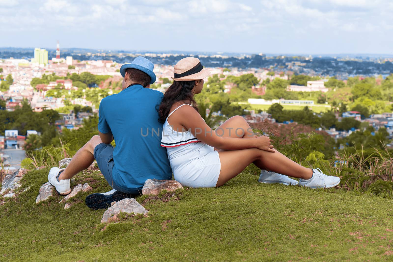 A couple of young people wearing hat sitting on the floor looking towards a city from a hill