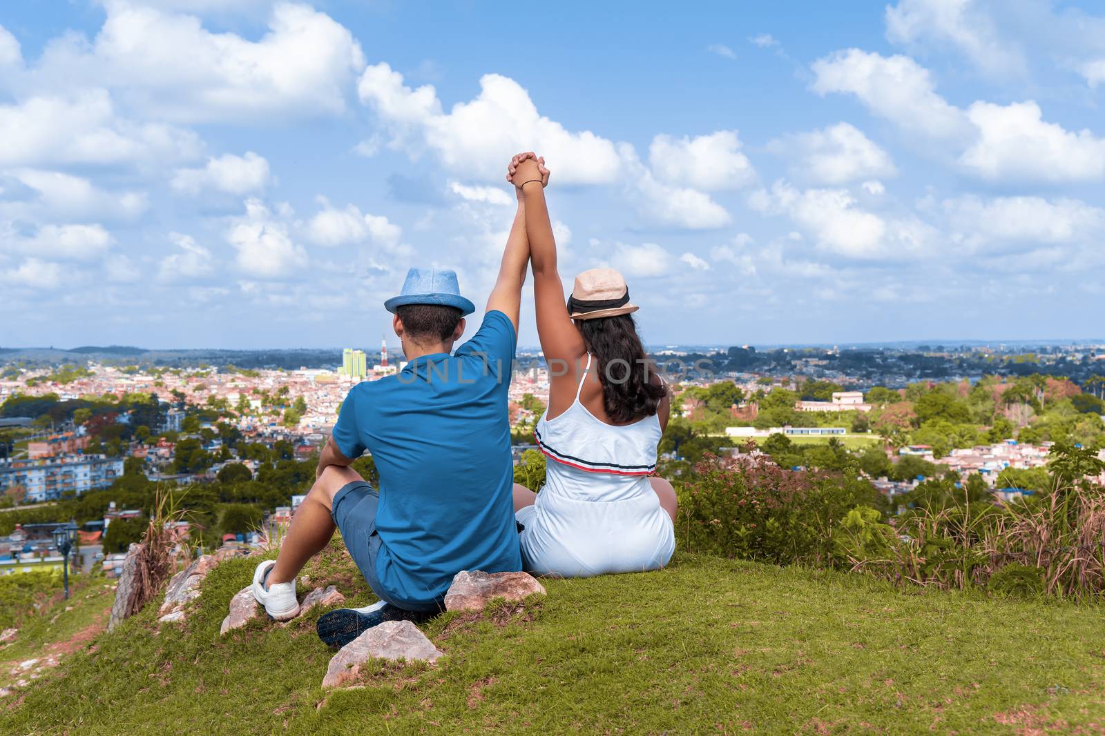 A couple of young people wearing hat with their arms up sitting on the floor looking towards a city from a hill