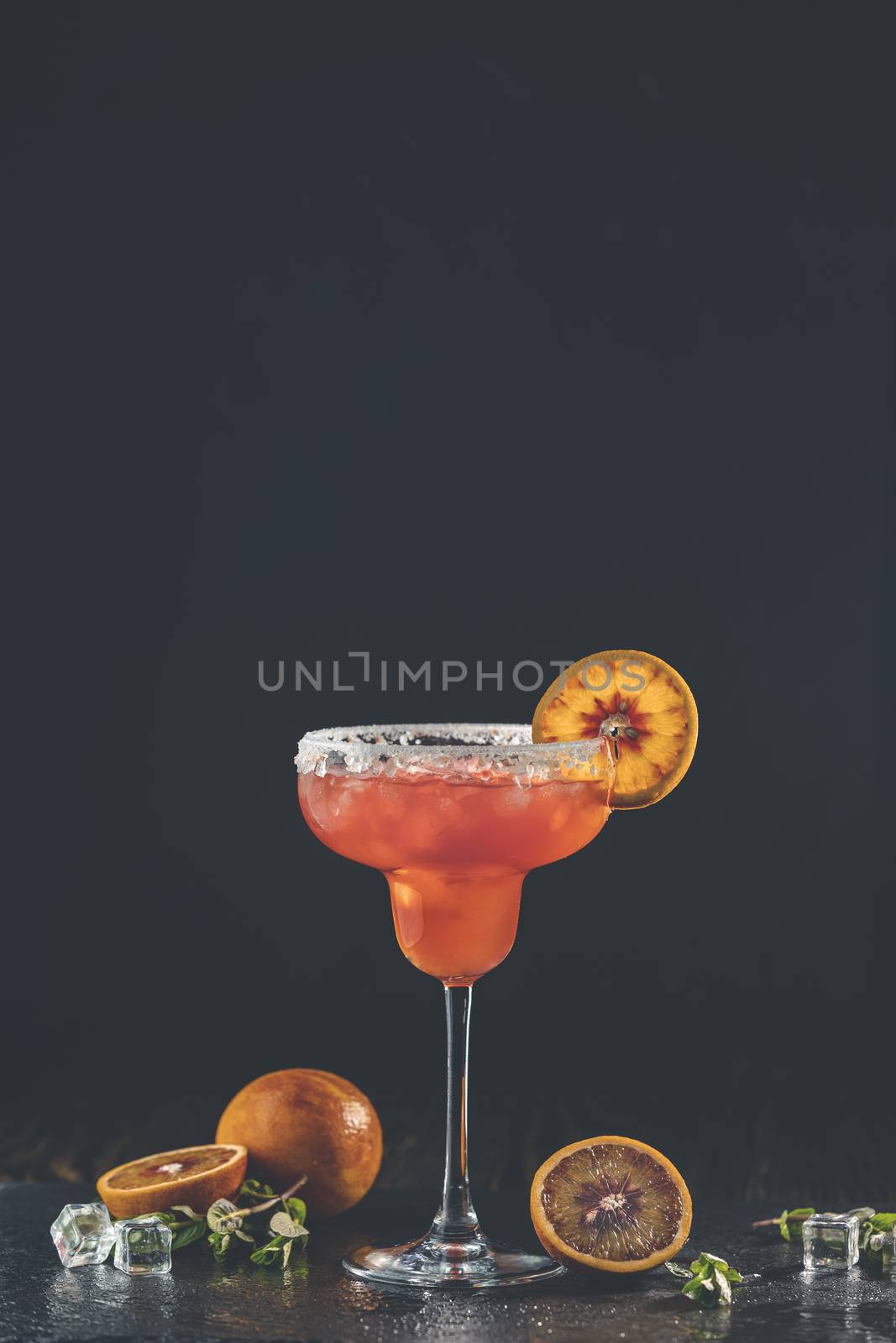 Red orange margarita cocktail with tequila, triple sec, orange juice, crushed ice and some salt on the rim of a glass, decorated with a citrus slice  Shallow depth of the field Dark stone background