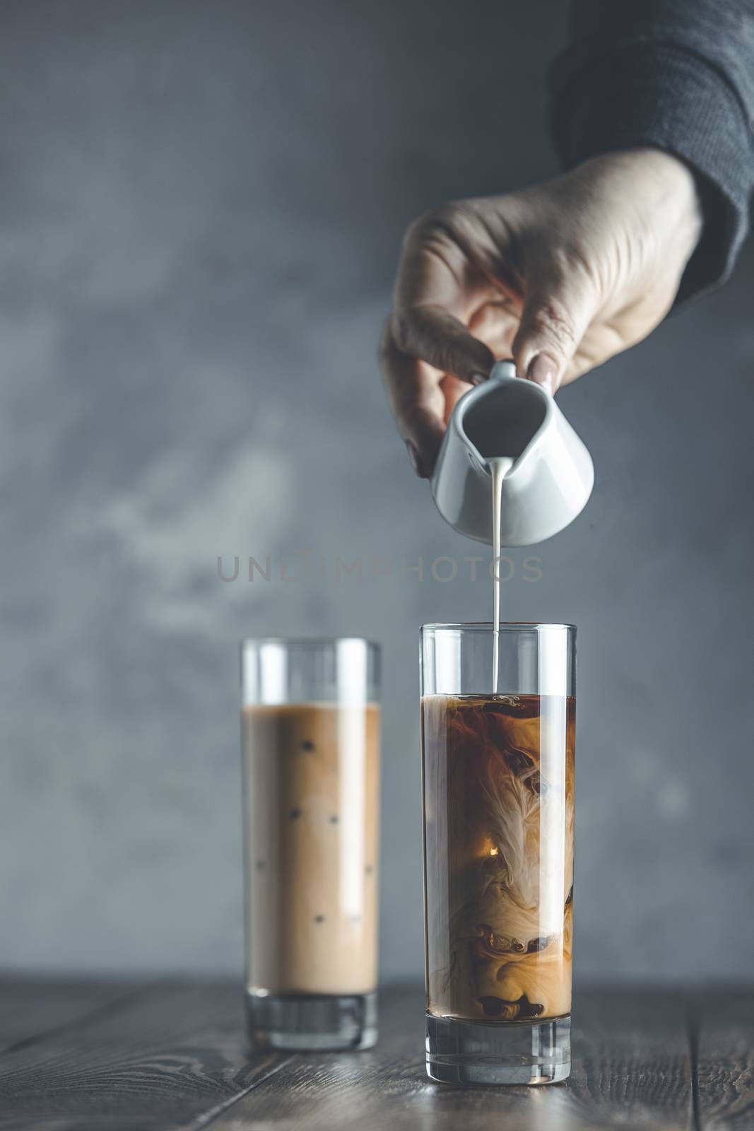 Women hand is pouring homemade sour cream from small jar to glass with brew cold coffee and ice. Cold summer drink on a dark wooden table and gray background with copy space.