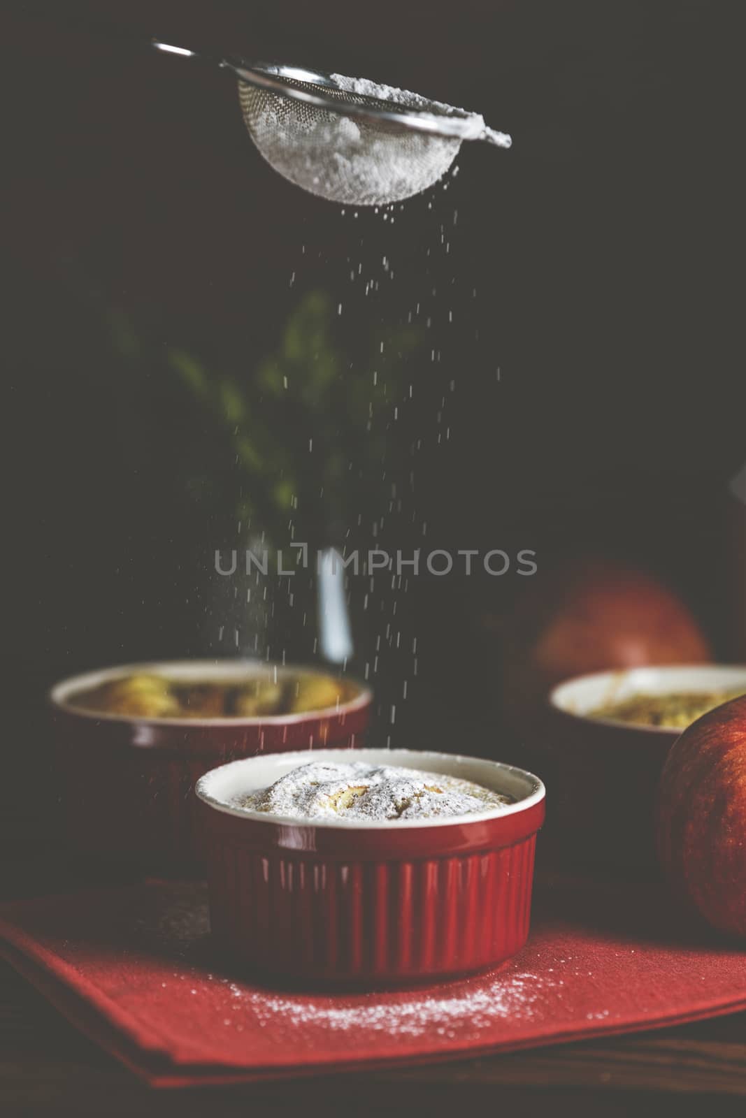 Sifting powdered sugar by sieve over apple pie in ceramic baking molds ramekin on dark wooden table. Close up, shallow depth of the field.