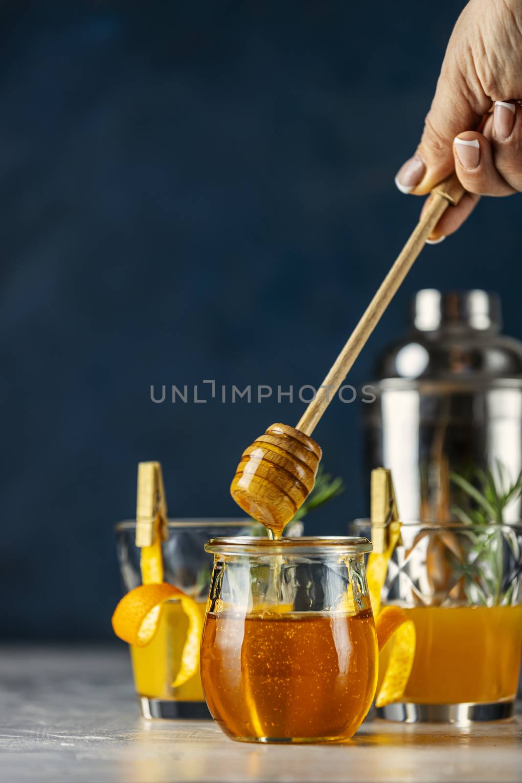 Woman hand holds a spoon for honey over jar in front of two glasses of honey bourbon cocktail with rosemary syrup or homemade whiskey sour with orange  peel and rosemary decoration and bartender tools