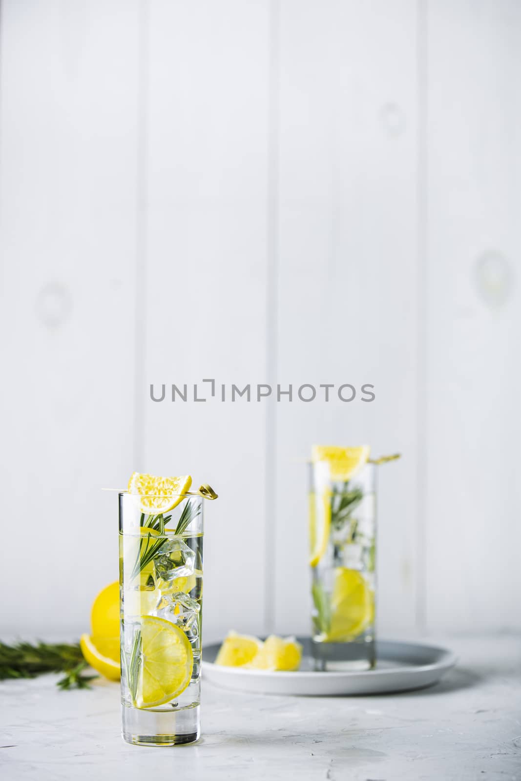 Two glasses of refreshing lemon lime drink with ice cubes in glass goblets against a light gray background. Summer fresh lemon soda cocktail with rosemary, selective focus