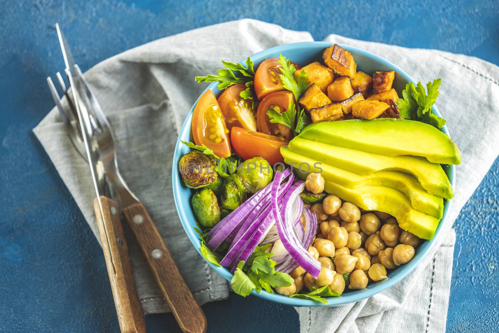Healthy food. Healthy vegan lunch bowl of fried sweet potato, chickpeas, red onion, avocado, tomatoes, and brussels sprouts. Top view, classic blue concrete surface, flat lay.