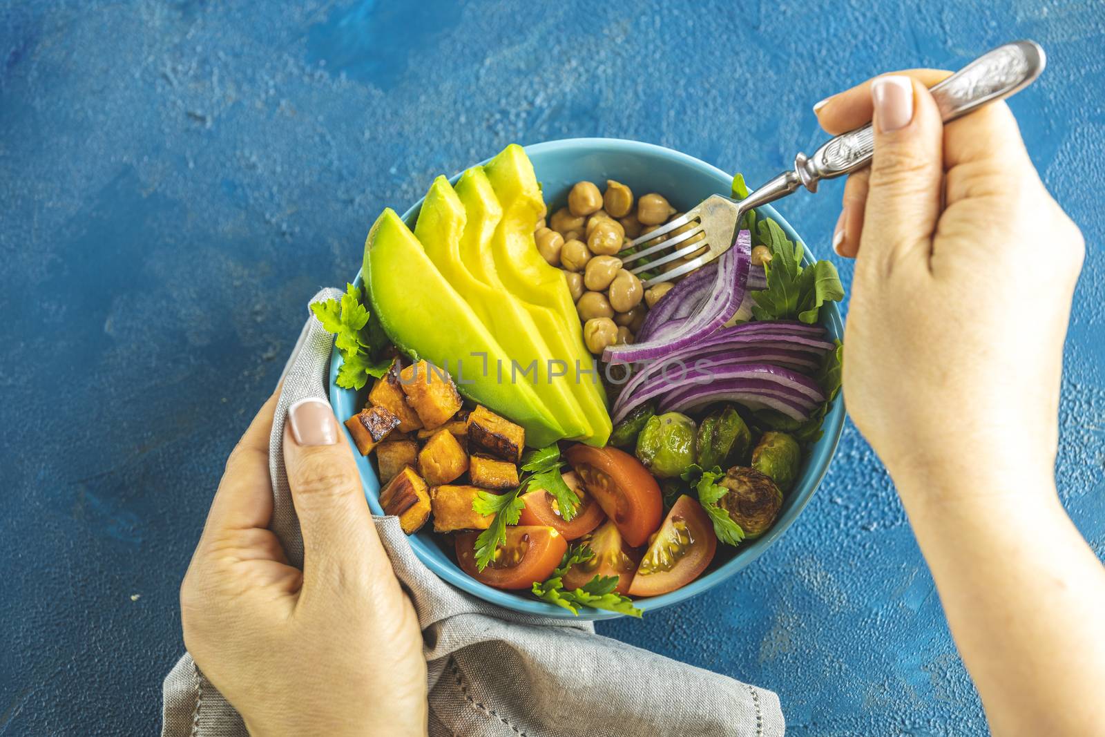Woman is eating healthy lunch. Vegan lunch bowl of fried sweet potato, chickpeas, red onion, avocado, tomatoes, and brussels sprouts. Top view, flat lay. Clean and balanced healthy food concept