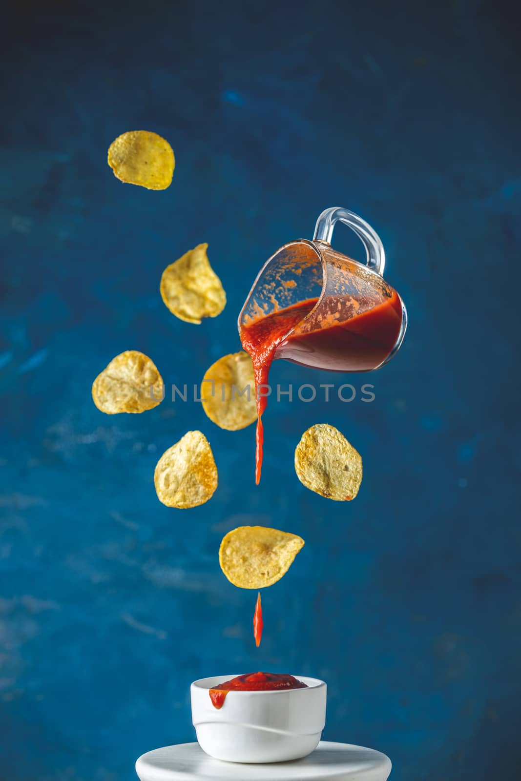 Sauce in glass jar and tasty potato chips falling into blow with tomato ketchup frozen in the air. Pouring homemade natural canned hot tomato sauce from small jar to white ceramic pot. Color of 2020