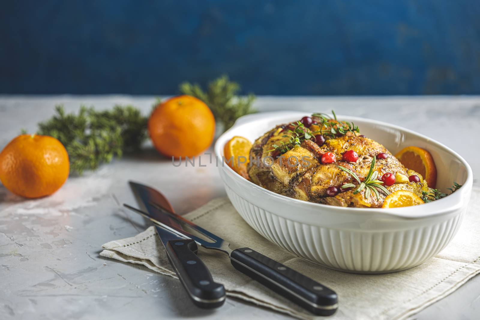 Roasted pork in white dish, christmas baked ham with cranberries, tangerines, thyme, rosemary, garlic on light table surface, close up.