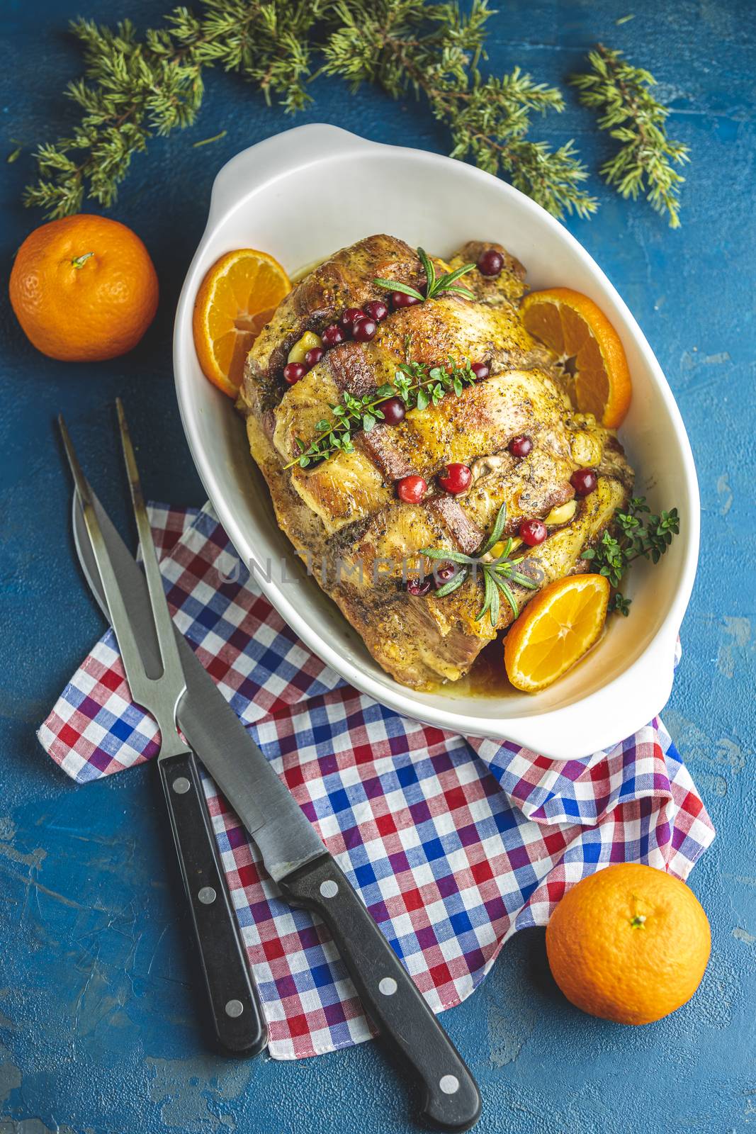 Roasted pork in white dish, christmas baked ham with cranberries, tangerines, thyme, rosemary, garlic on light table surface, close up.