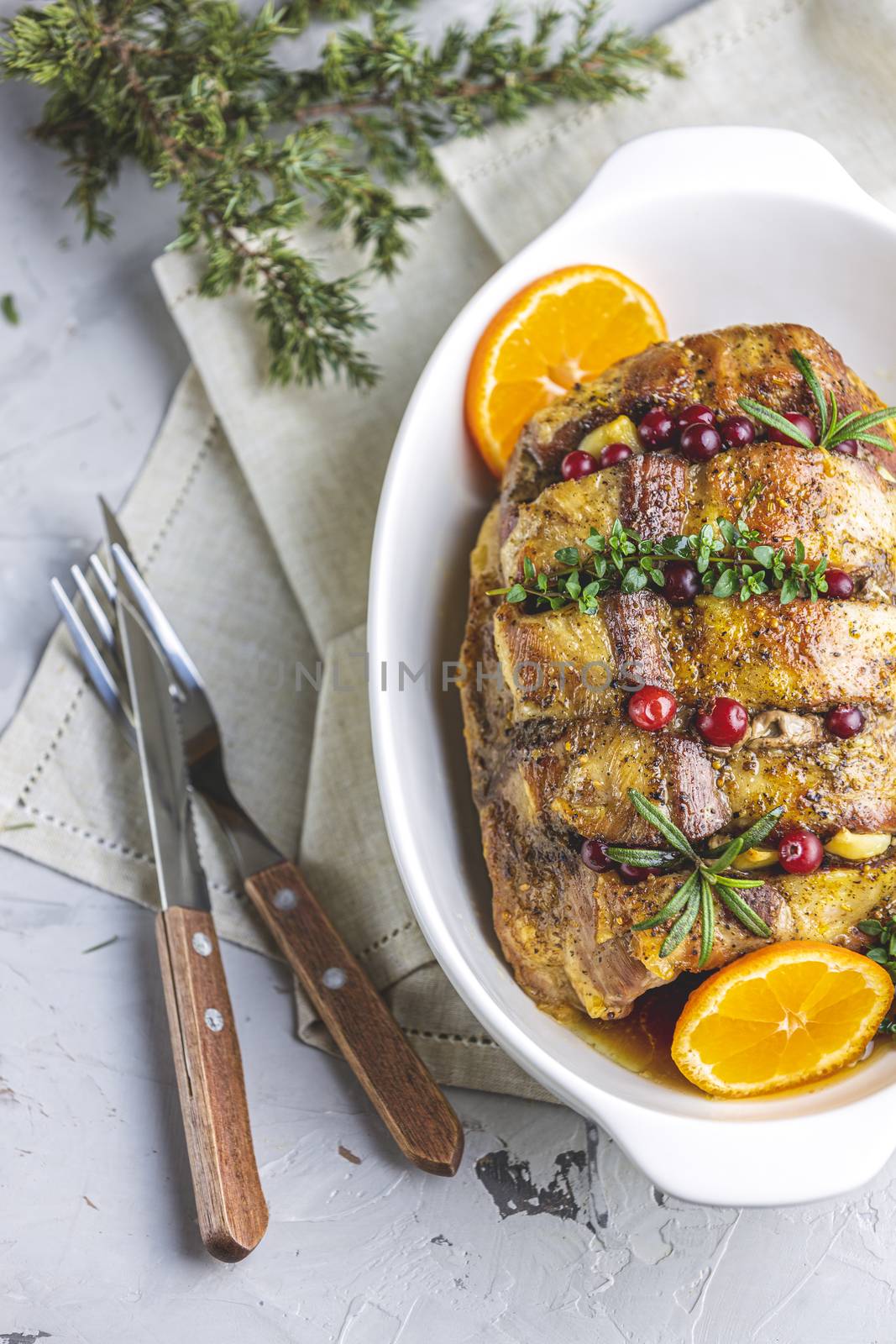 Roasted pork in white dish, christmas baked ham with cranberries, tangerines, thyme, rosemary, garlic on light table surface, close up.