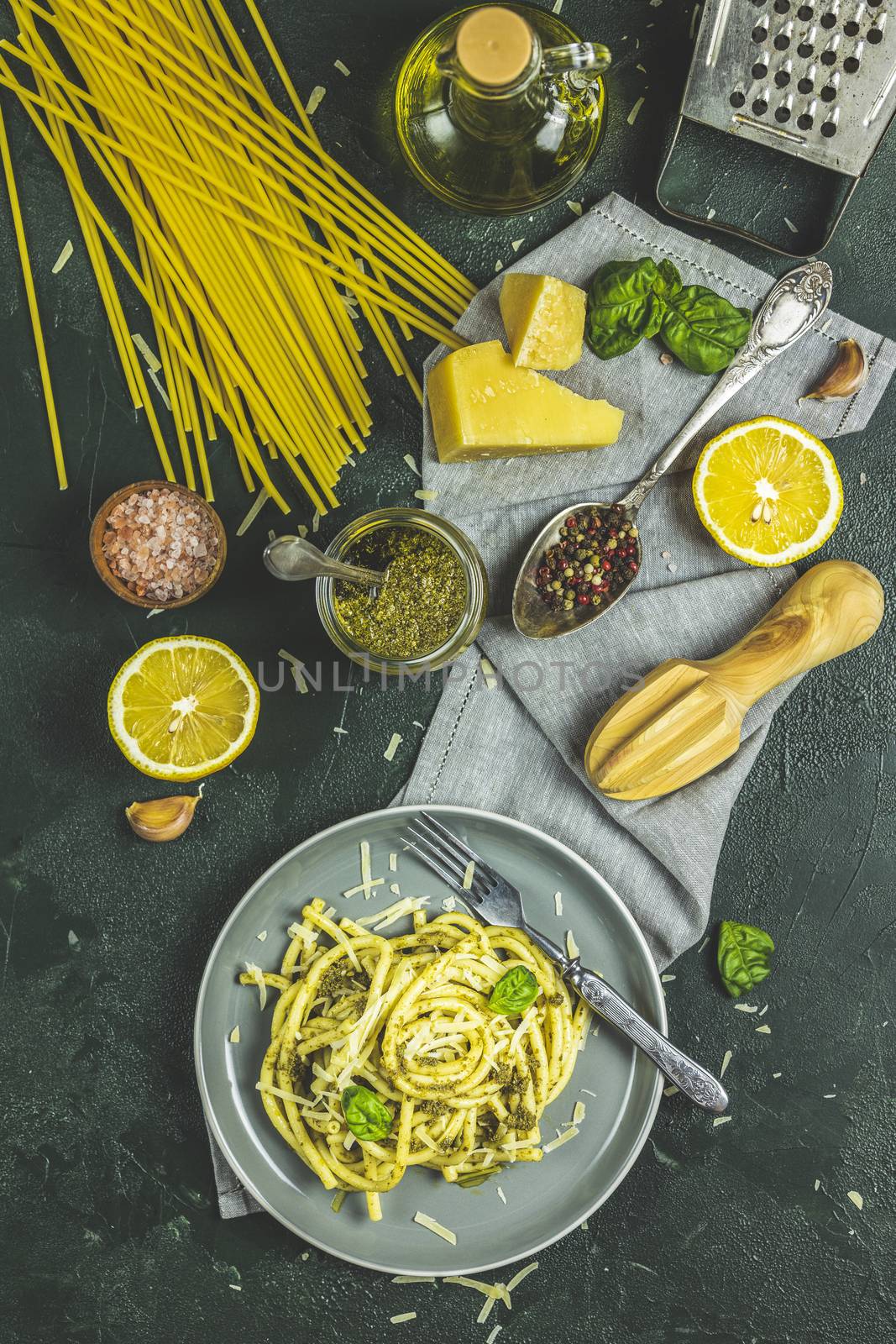 Spaghetti pasta bucatini with pesto sauce and parmesan. Italian traditional perciatelli pasta by genovese pesto sauce in gray dish served with ingredients. Flat lay on dark green surface.