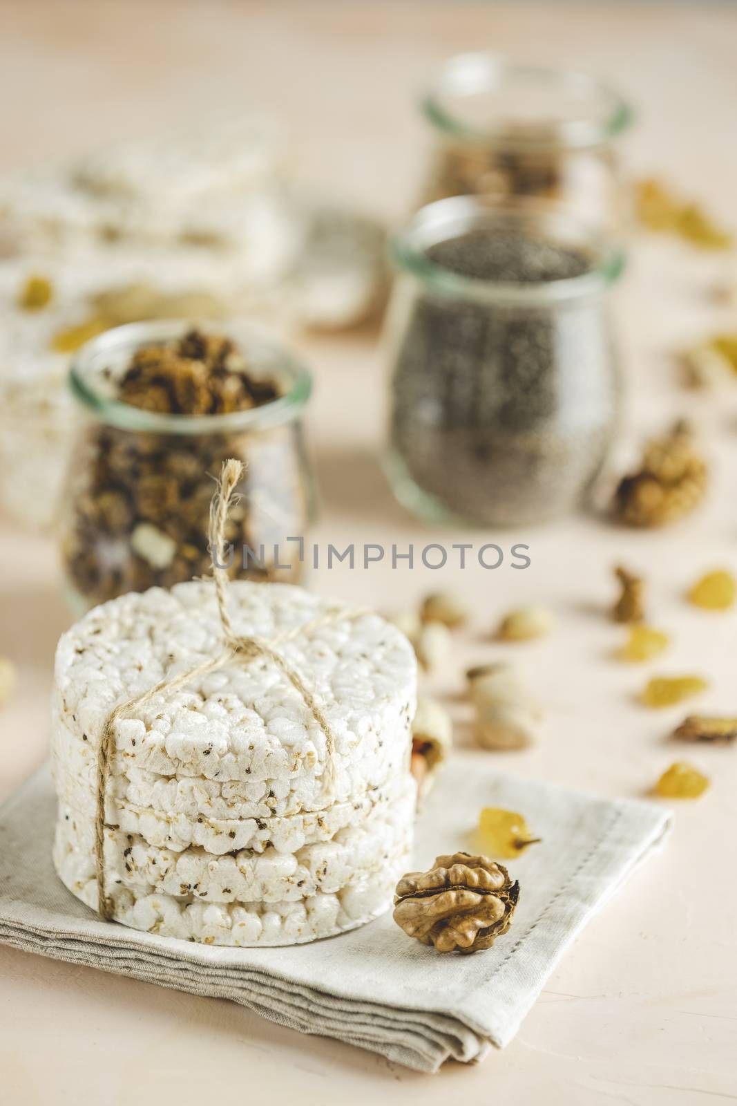 American puffed rice cakes. Healthy snacks with almonds, raisins, peanuts, pistachios in glass jars on light pink concrete surface.	