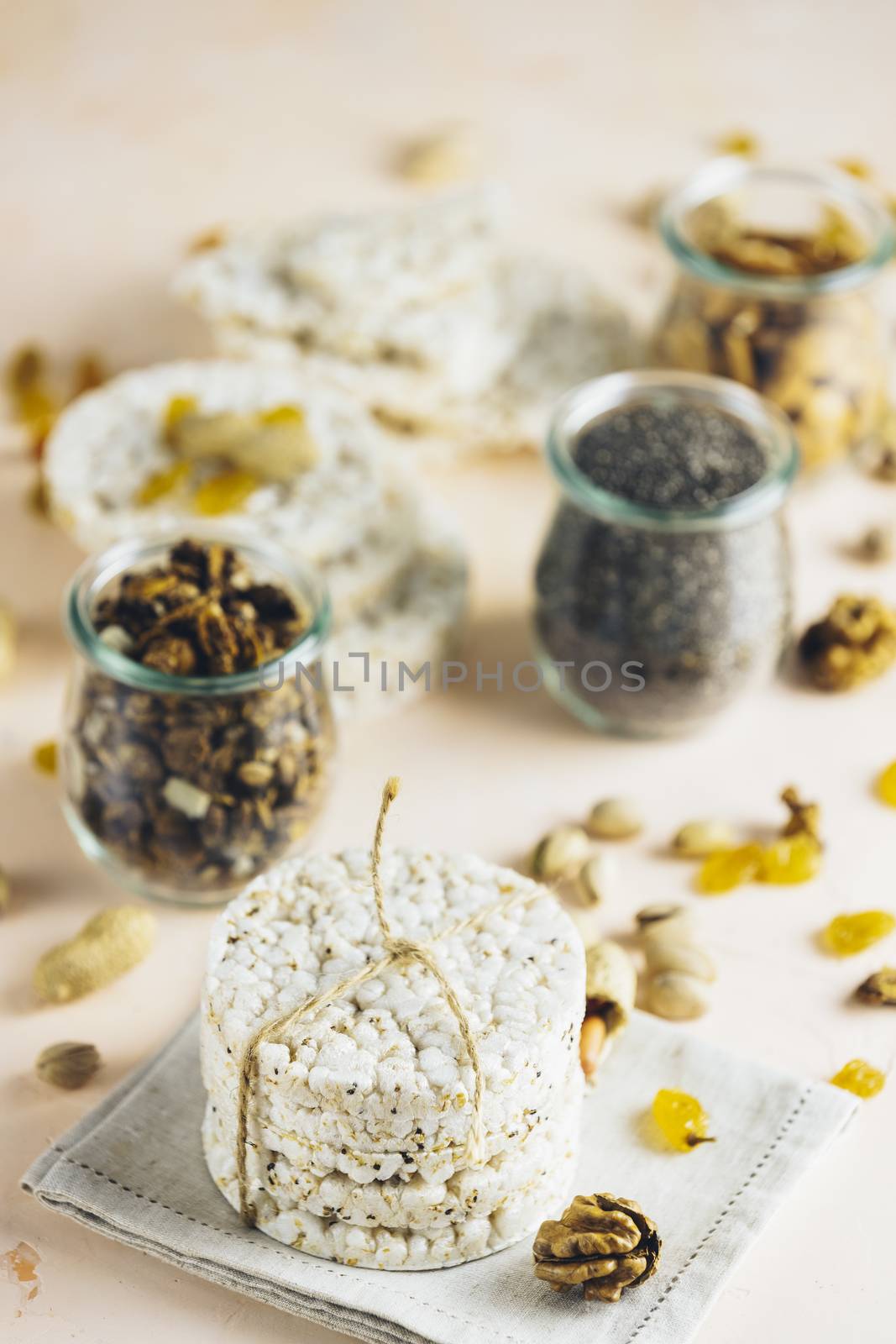 American puffed rice cakes. Healthy snacks with almonds, raisins, peanuts, pistachios in glass jars on light pink concrete surface.