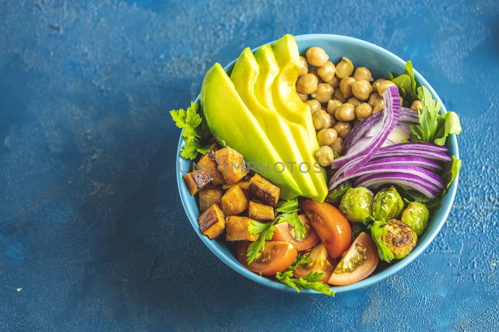 Healthy food. Healthy vegan lunch bowl of fried sweet potato, chickpeas, red onion, avocado, tomatoes, and brussels sprouts. Top view, classic blue concrete surface, flat lay
