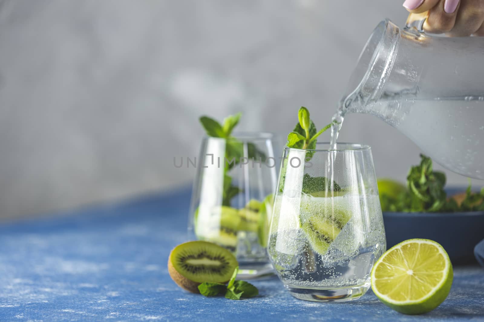 Women hand is pouring detox water, mojito or martini tonic cocktail from glass jar to glass with kiwi, lime, ice and mint. Summer fresh lime soda cocktail, selective focus.