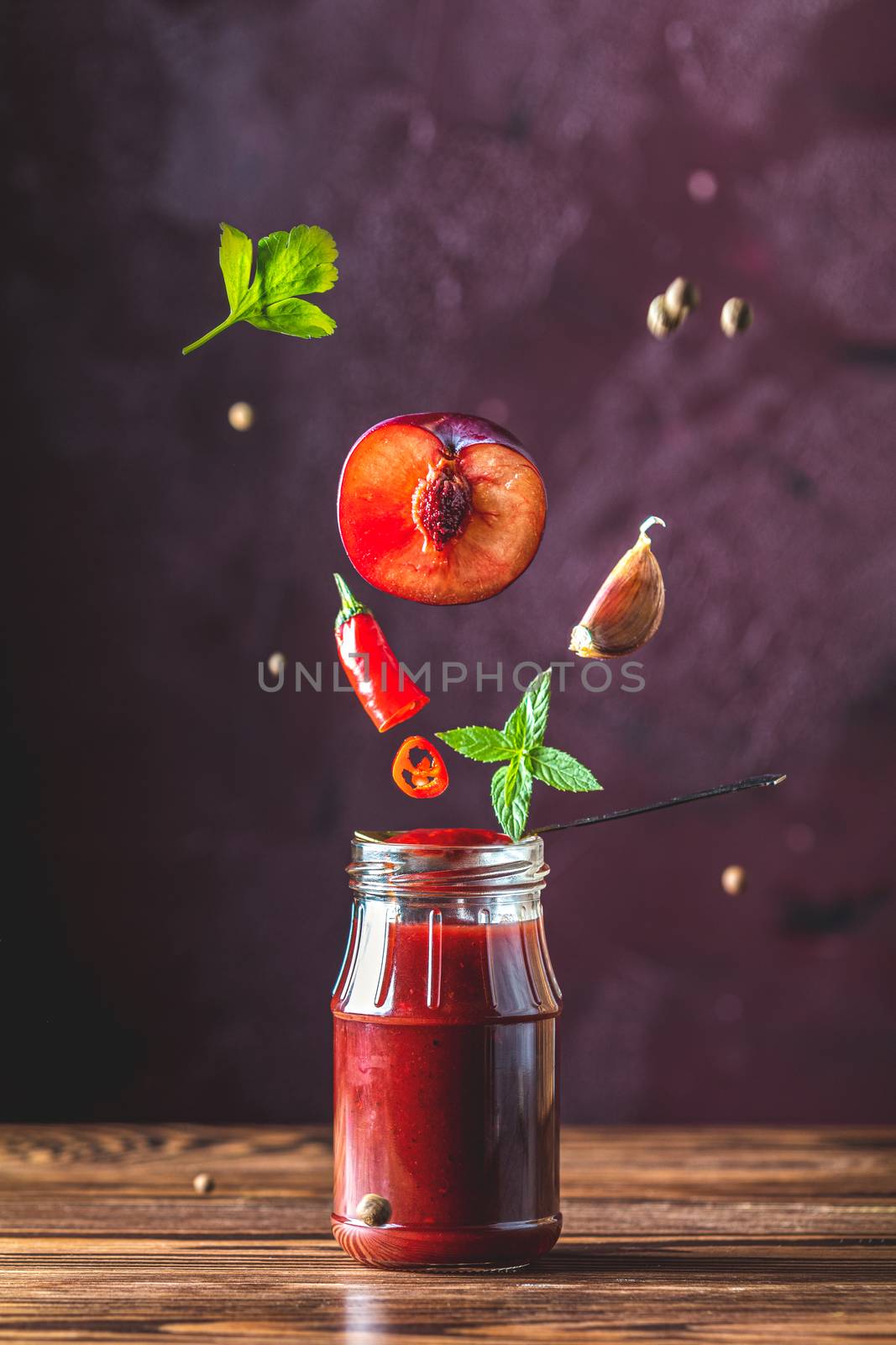 Sauce components frozen in the air. Homemade DIY natural canned hot plum sauce chutney with chilli or tkemali in glass jar standing on wooden table with flying ingredients, selective focus