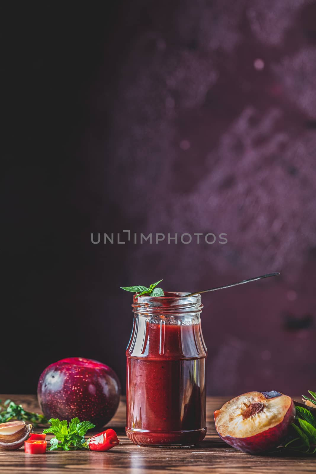 Homemade DIY natural canned hot plum sauce chutney with chilli or tkemali in glass jar standing on wooden table with ingredients — plum, salt, pepper, herbs, selective focus