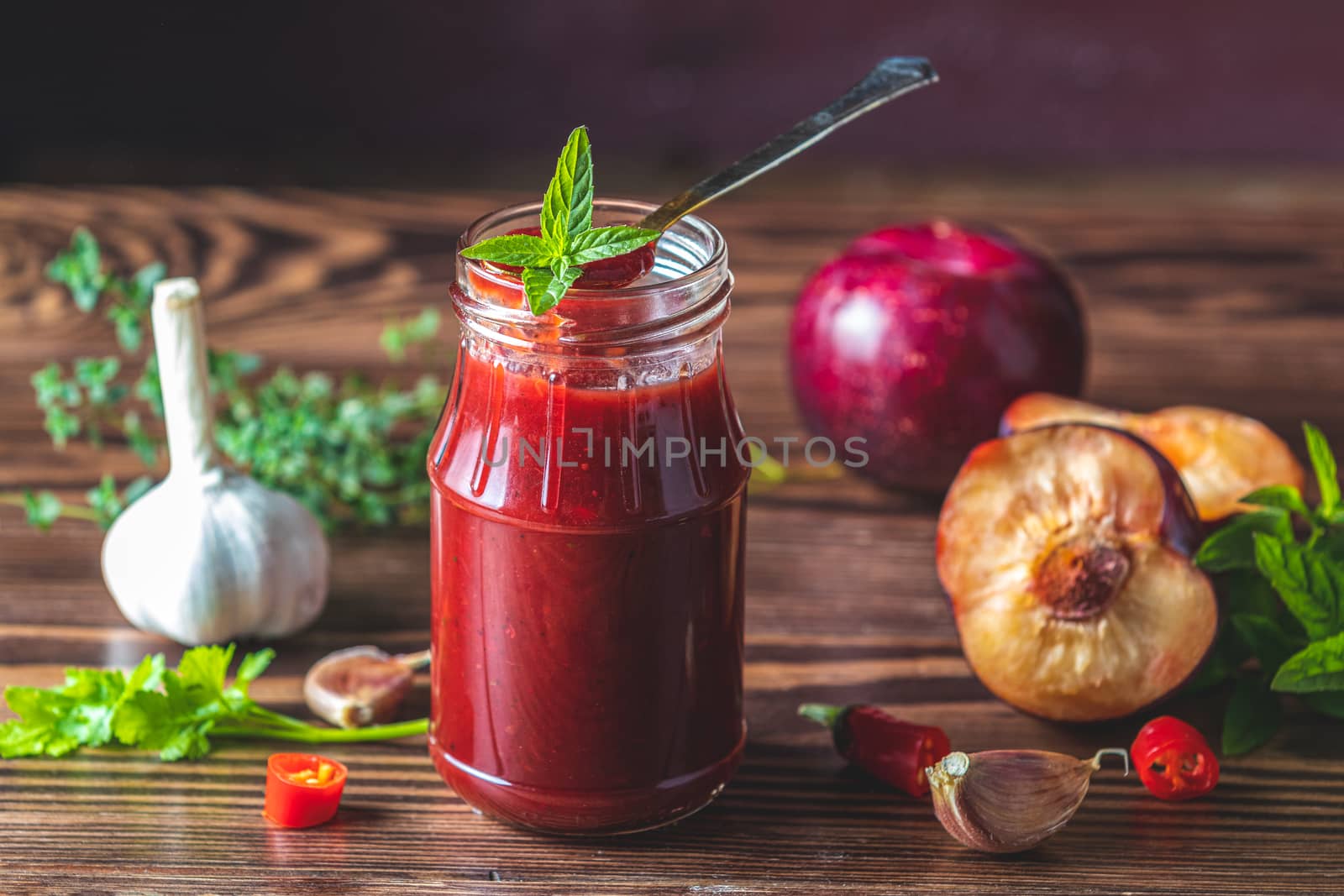 Homemade DIY natural canned hot plum sauce chutney with chilli or tkemali in glass jar standing on wooden table with ingredients — plum, salt, pepper,  herbs, selective focus